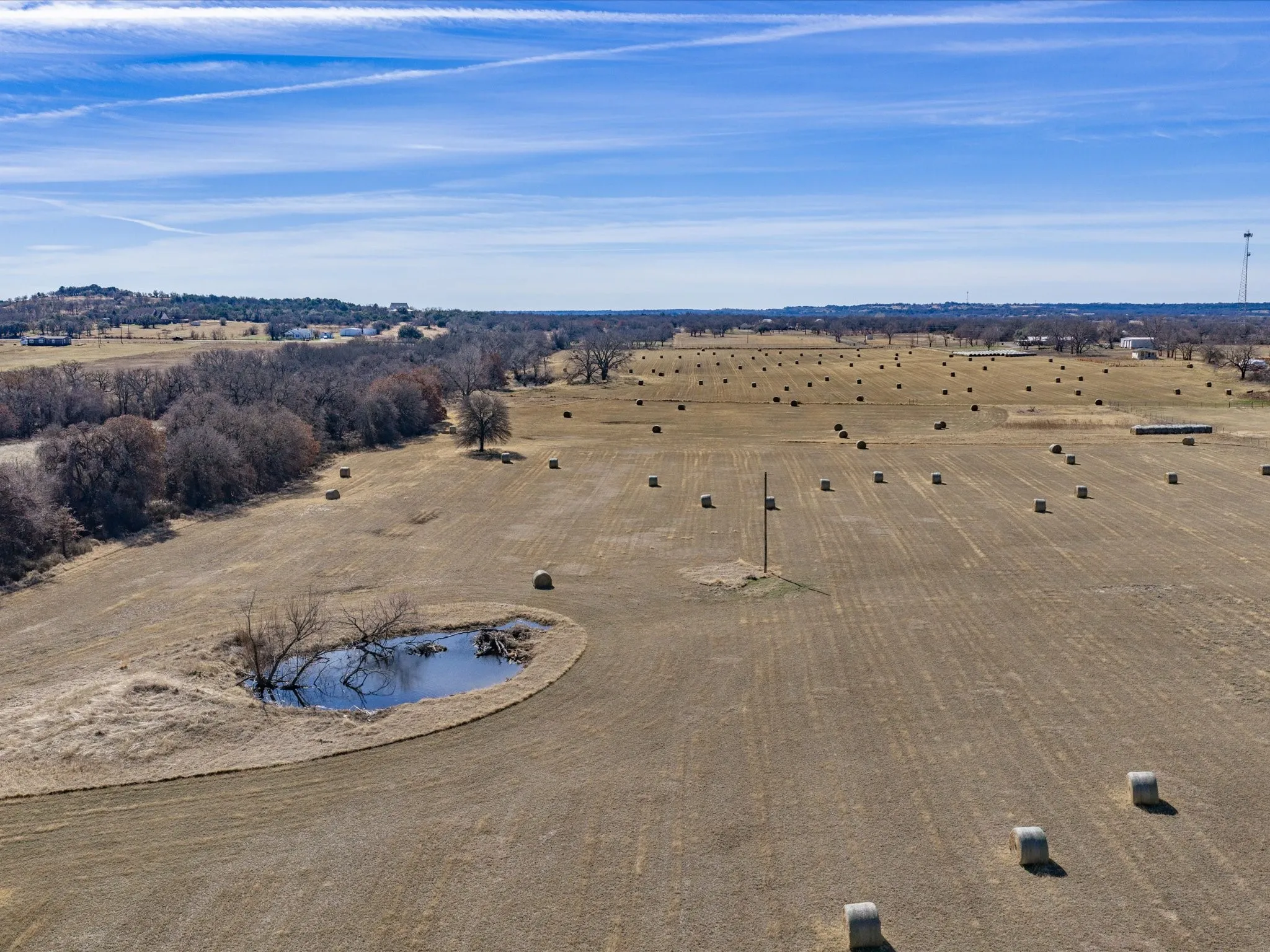Expansive agricultural land featuring a natural pond, scattered hay bales, and perimeter tree lines