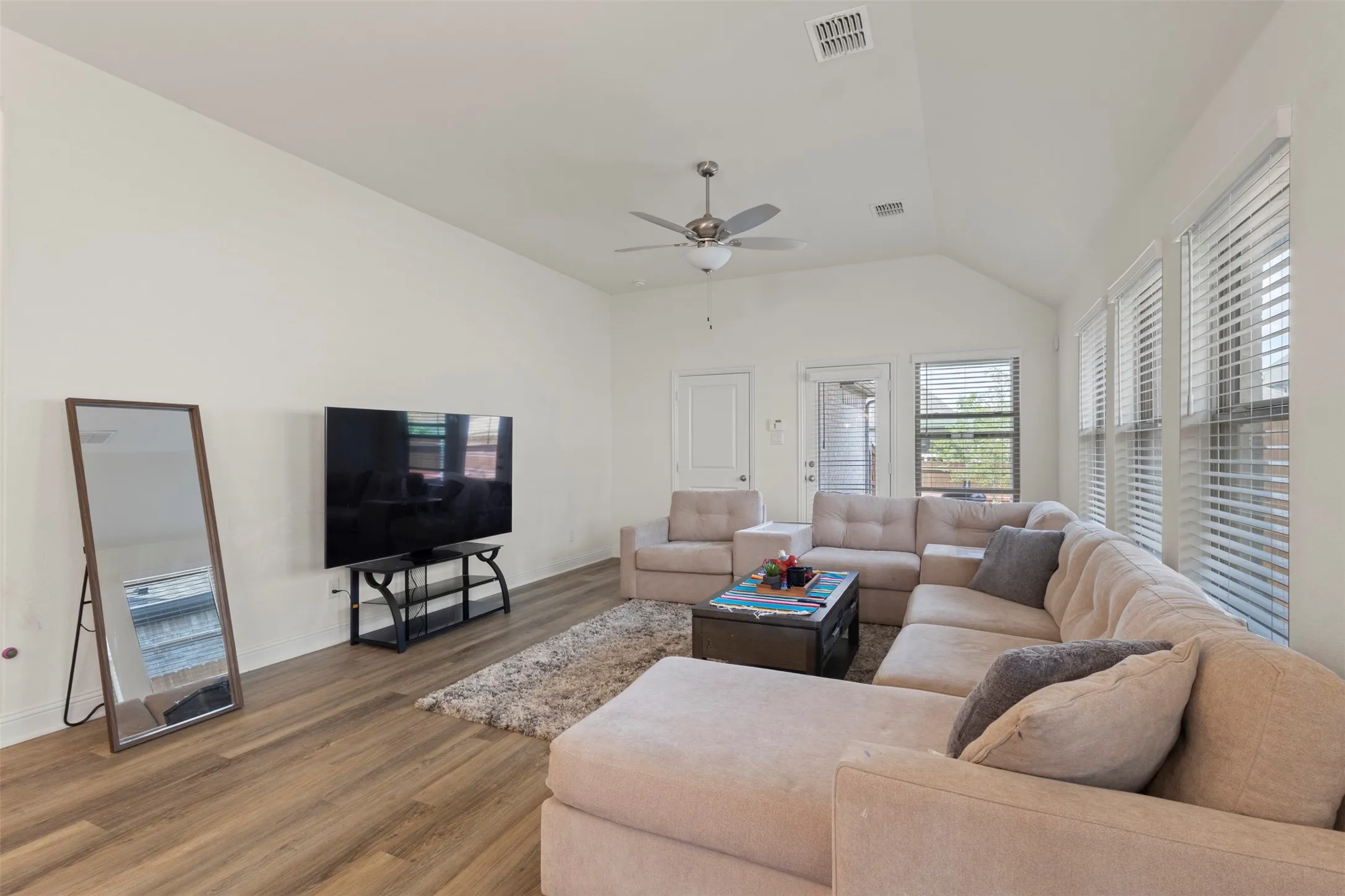 Vaulted ceilings, wood-finish flooring, ceiling fan, multiple windows with blinds, and a glass-paneled door