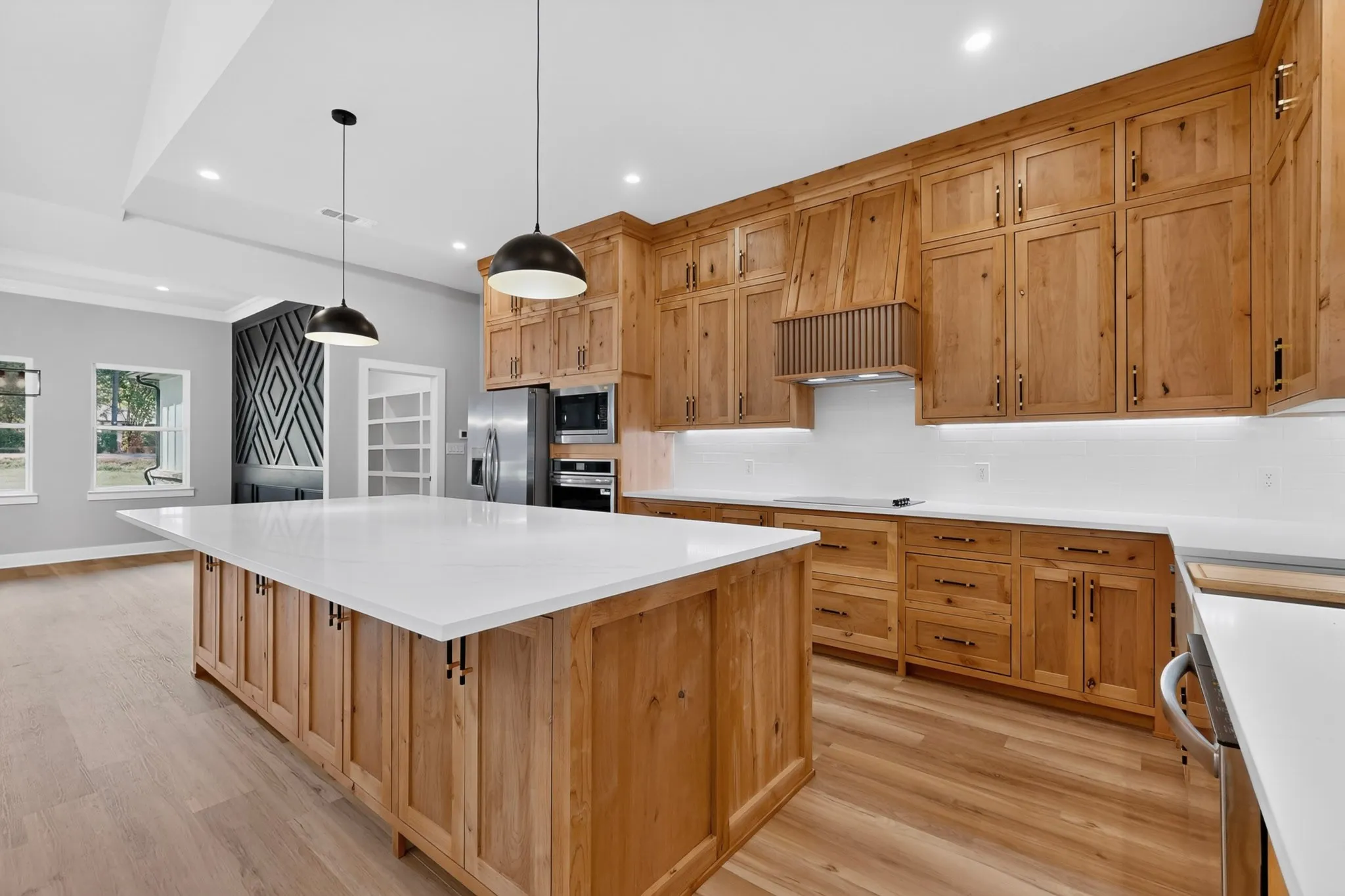Spacious kitchen featuring a large central island with a white countertop, extensive wood-finish cabinetry, stainless steel appliances, and pendant lighting