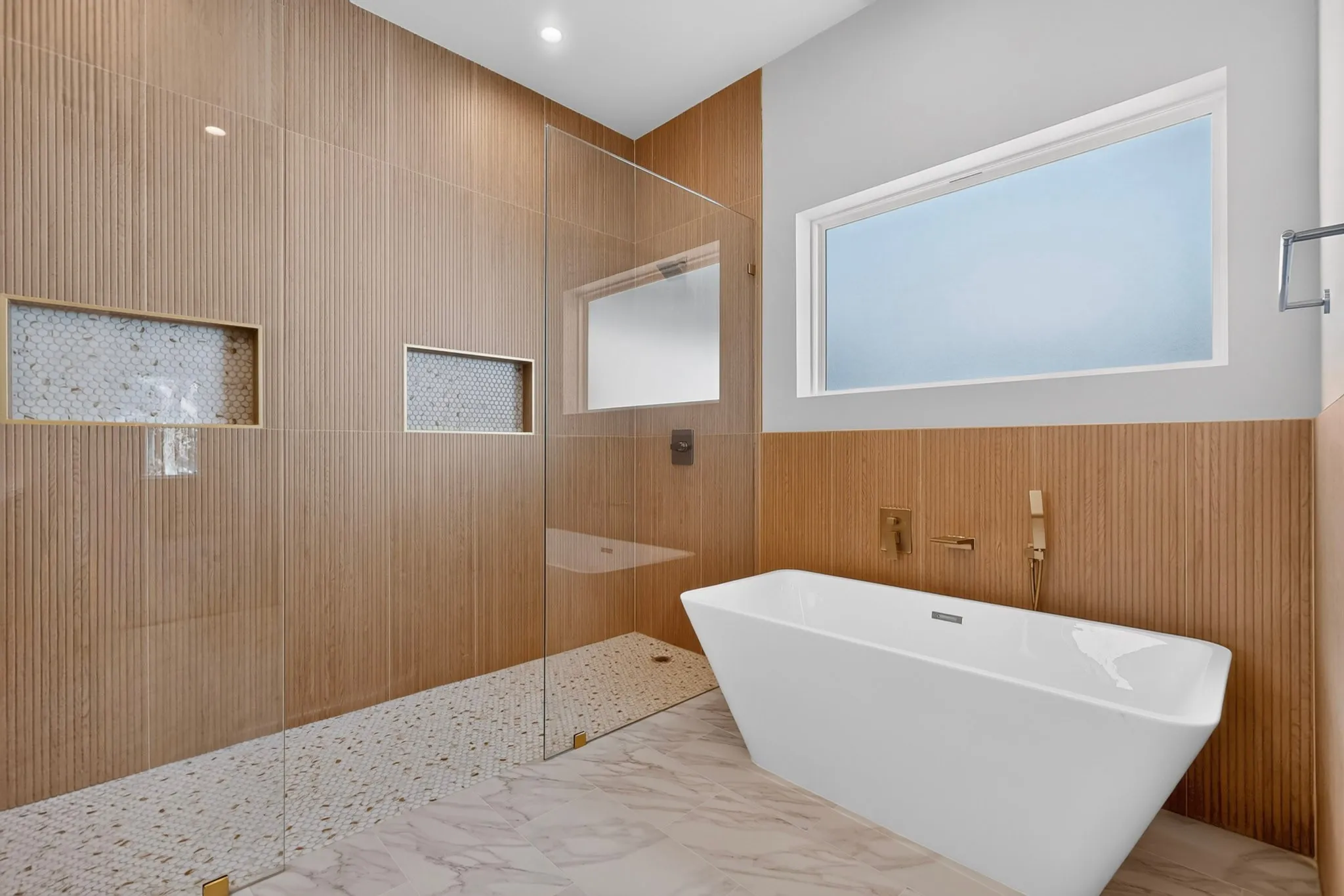 Freestanding soaking tub, walk-in shower with clear glass enclosure, textured tile walls, two recessed shower niches, and large format floor tiling