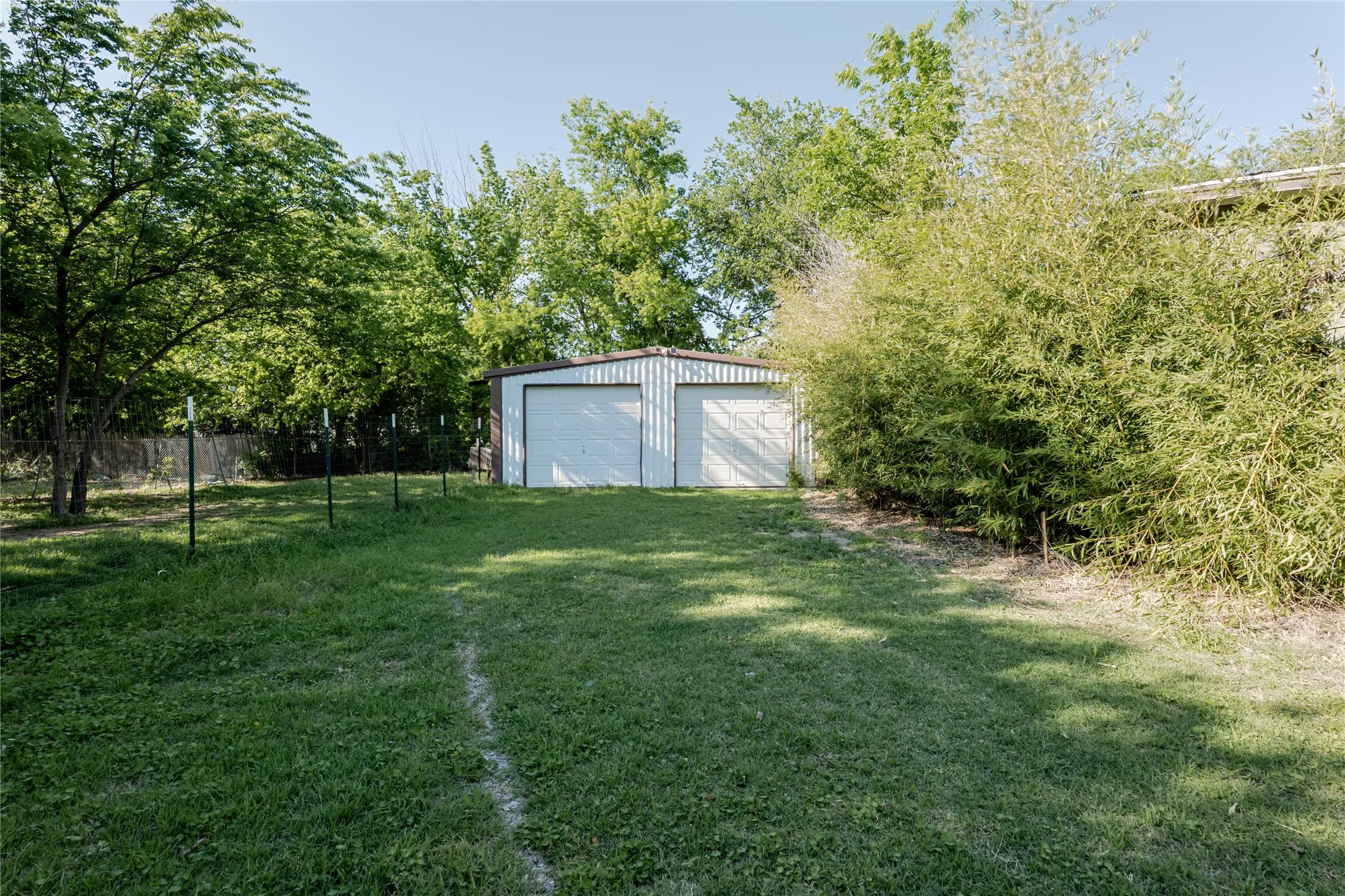Detached two-car metal garage building with dual roll-up doors, surrounded by mature trees and lush green landscaping