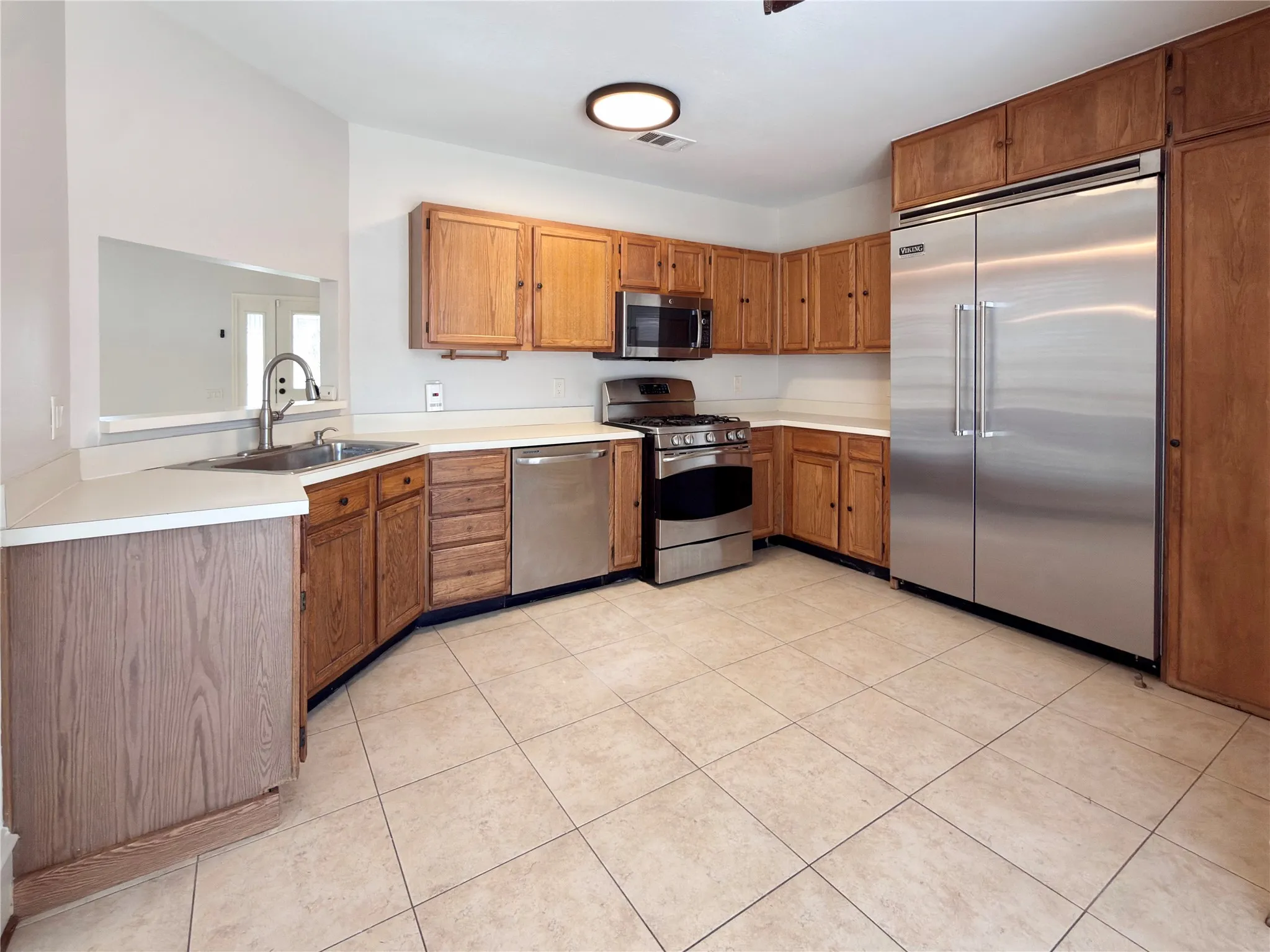 Kitchen featuring wood cabinetry, stainless steel appliances, a stainless steel sink with a gooseneck faucet, light-colored countertops, and tiled flooring
