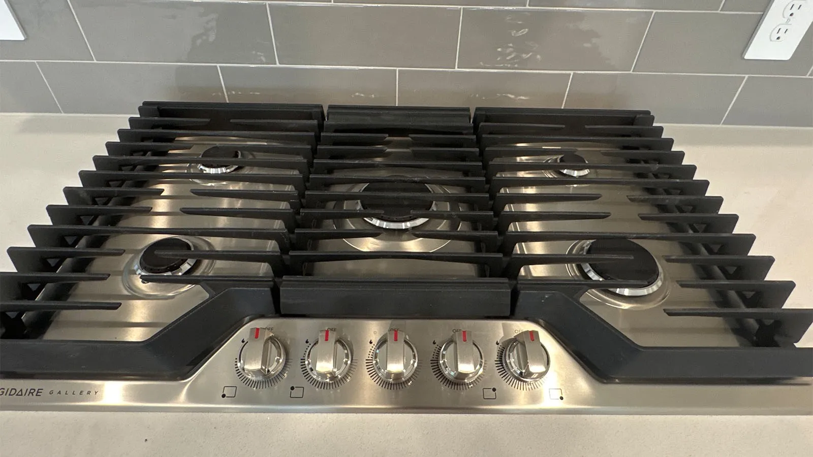 Stainless steel gas cooktop with five burners, cast iron grates, and a grey tile backsplash