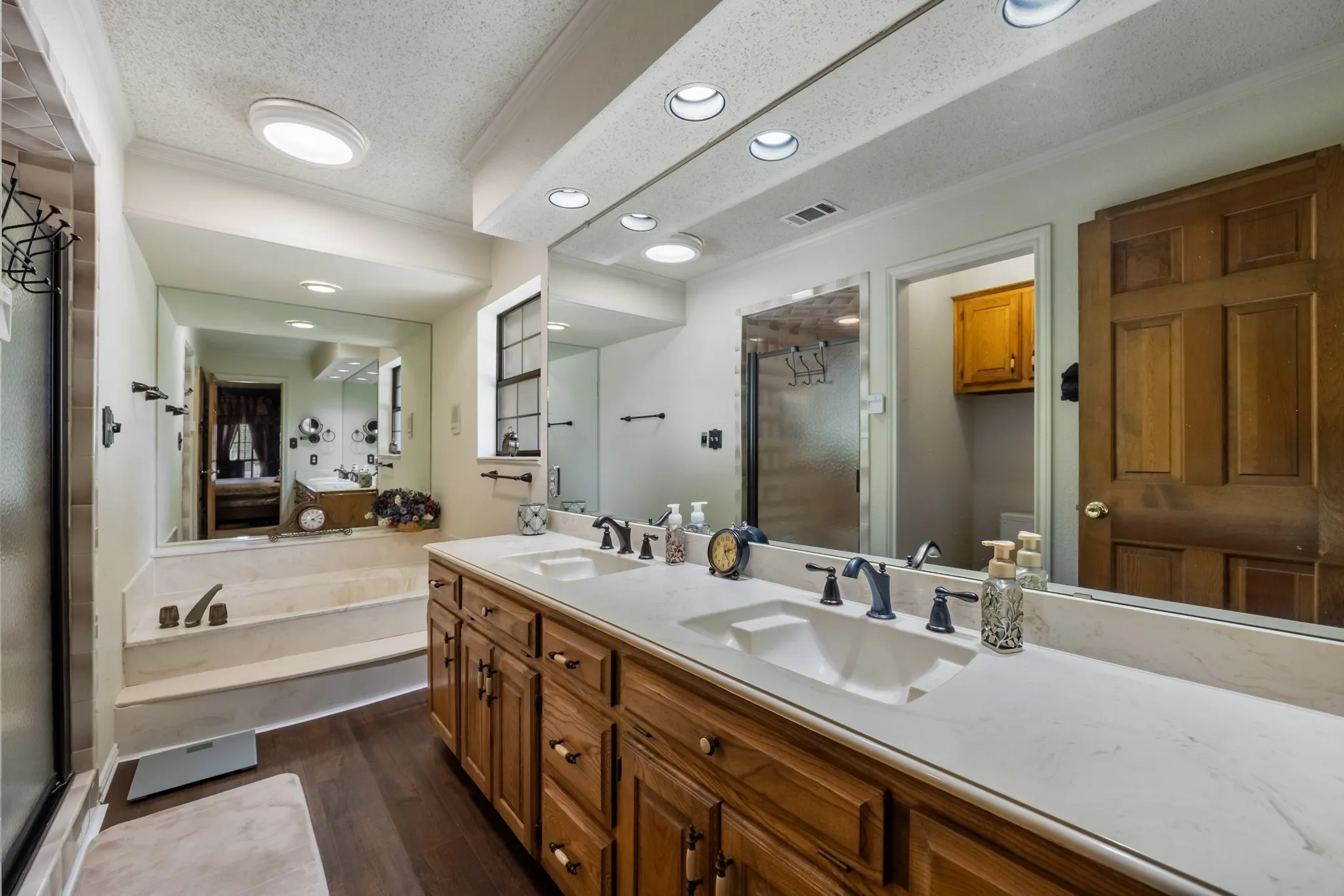 Dual vanity with integrated sinks, recessed lighting, wood-finish flooring, full-width mirror, and a built-in tub