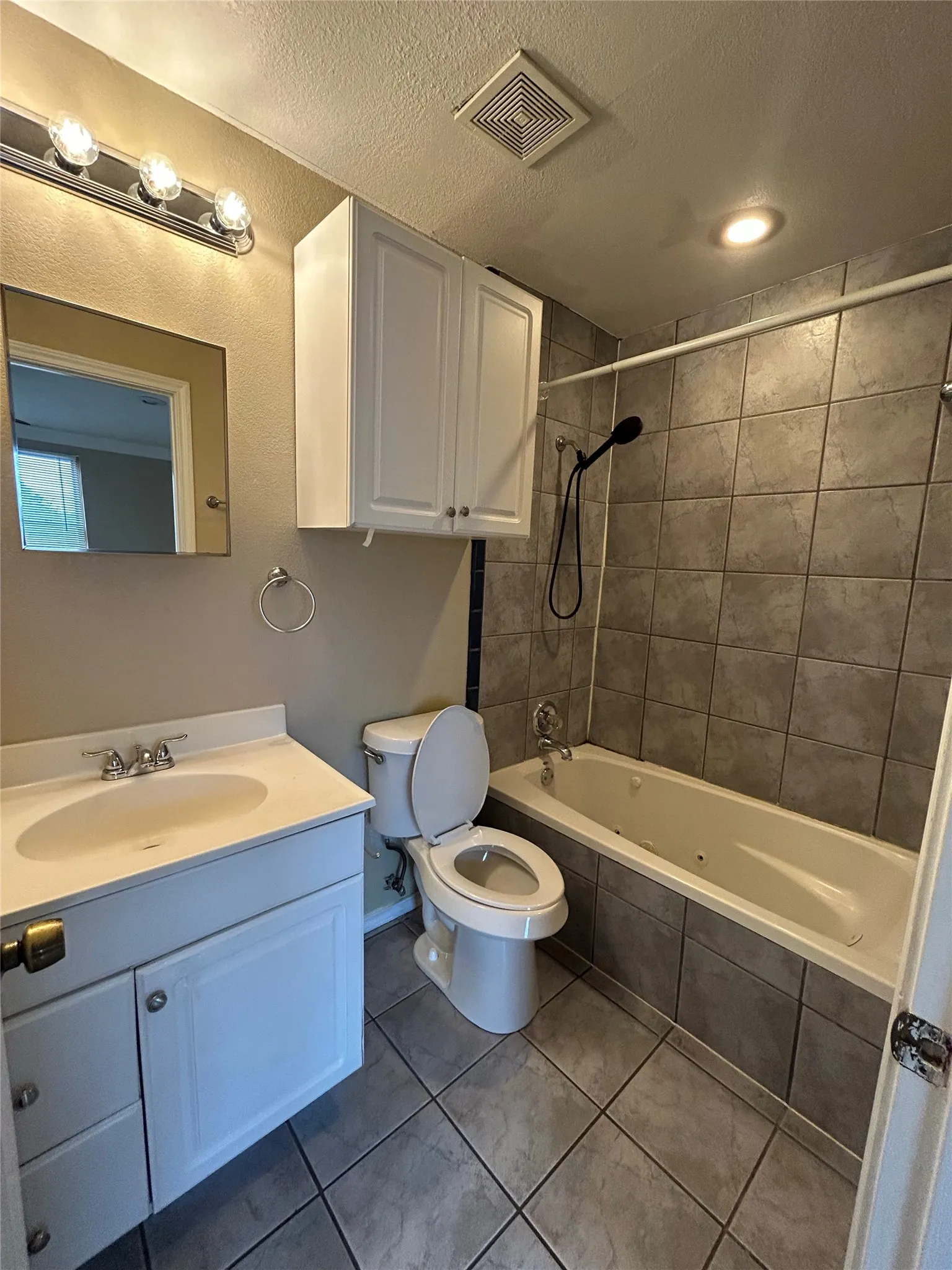 White vanity with integrated sink, overhead wall-mounted cabinetry, tile flooring, and a jetted tub with tiled surround