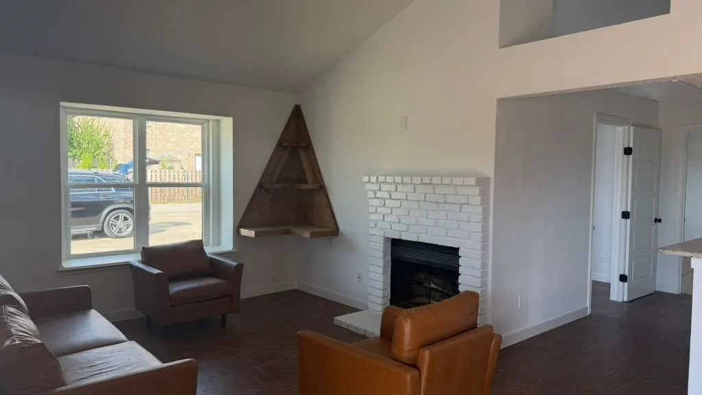 Vaulted ceiling, white brick fireplace, wood-finish flooring, built-in corner shelving, and an open doorway