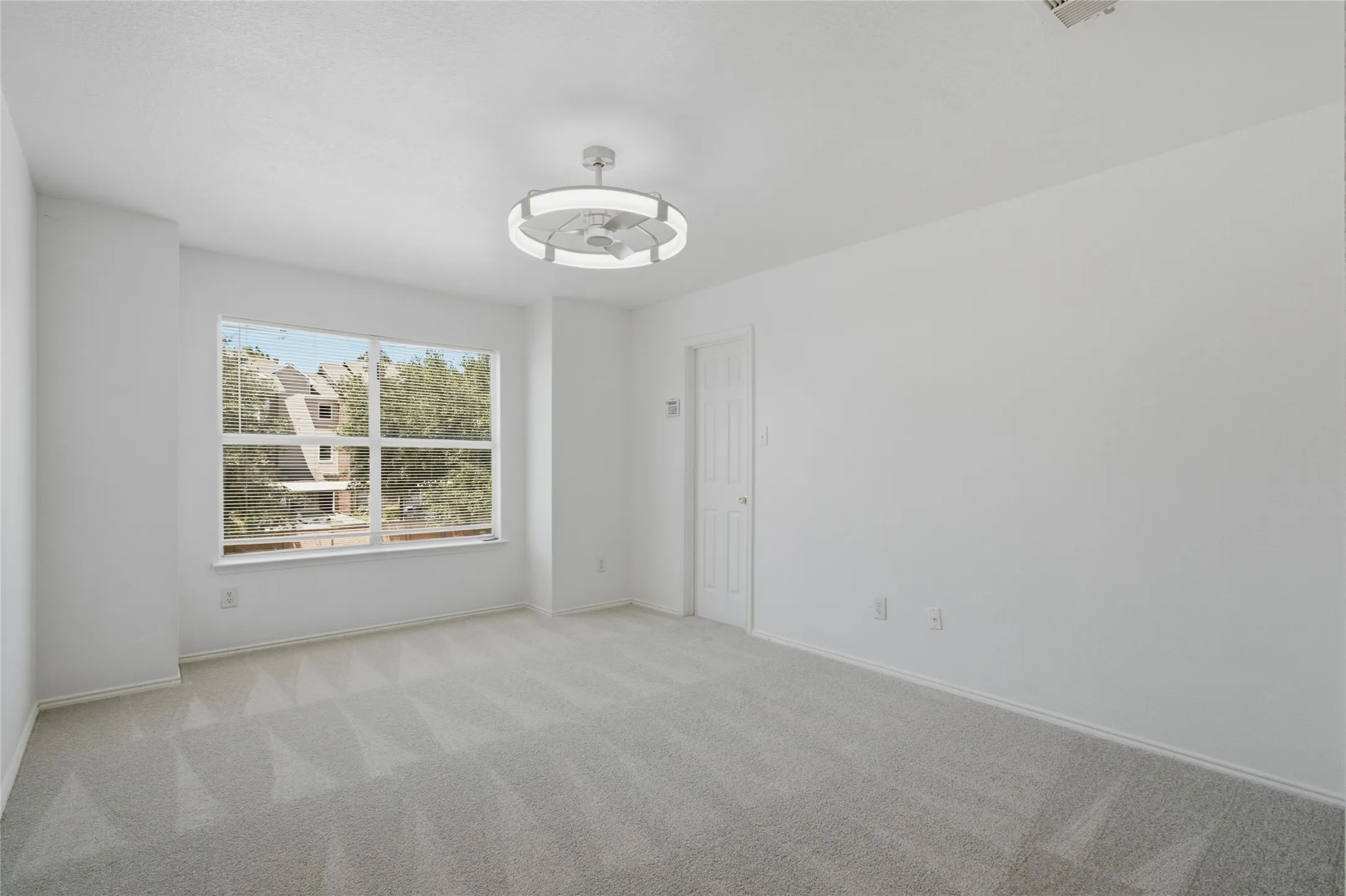 Spacious room featuring light gray patterned carpeting, white walls, a large multi-pane window, and a modern ceiling fan with integrated lighting