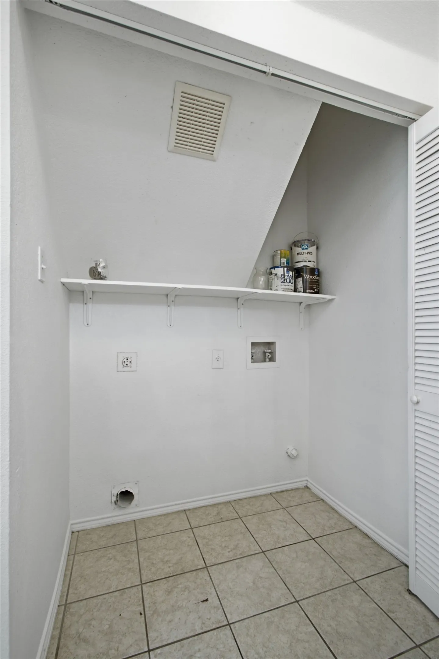 Laundry area featuring a tile floor, white shelving, an overhead vent, electrical outlets, and plumbing hookups