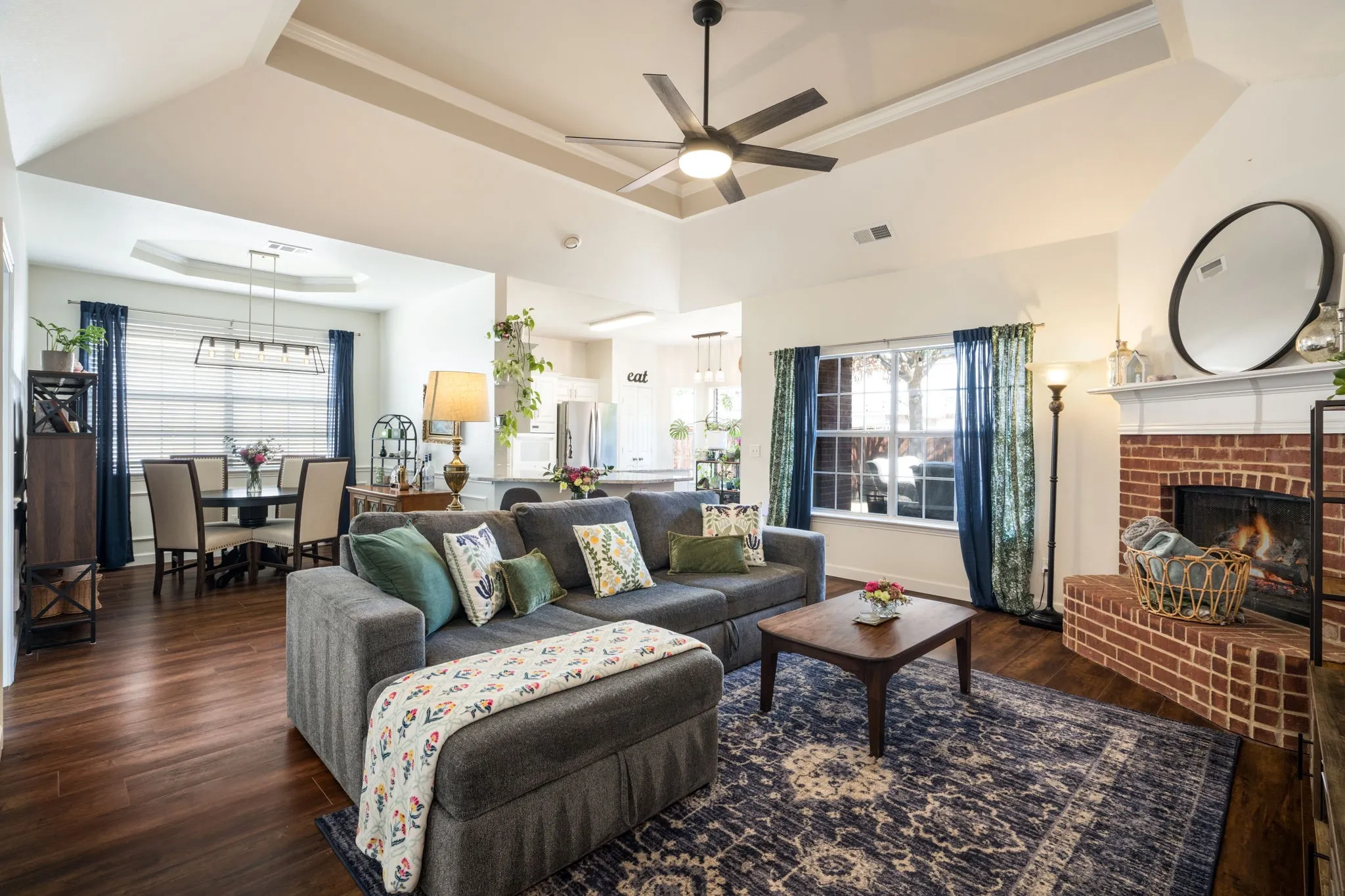 Tray ceiling with a ceiling fan, brick-surround fireplace with a white mantel, and wood-finish flooring