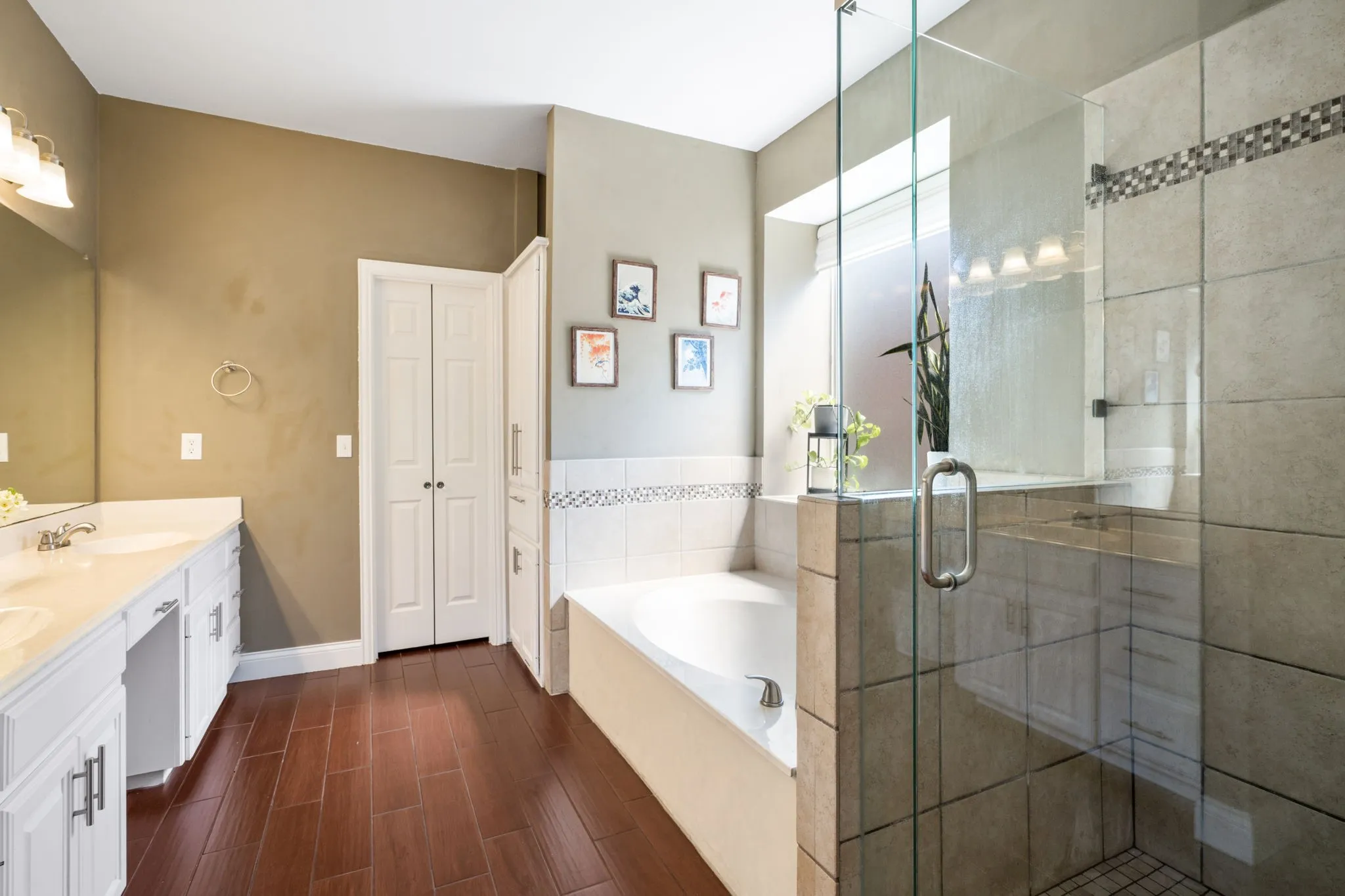 Double vanity with white cabinetry, wood-finish flooring, built-in soaking tub, glass-enclosed shower, and a built-in linen cabinet