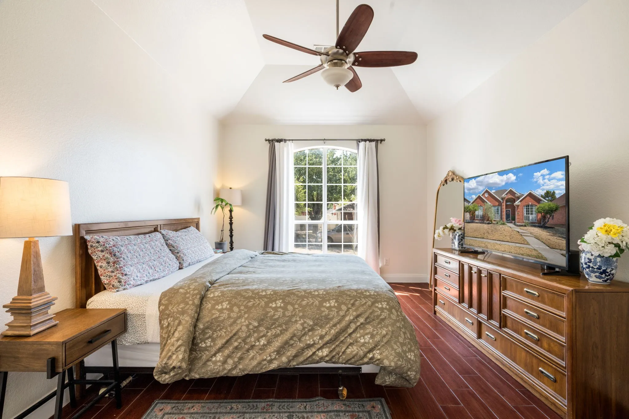 Vaulted ceiling with a ceiling fan, arched window with drapes, wood-finish flooring, and a built-in wood-finish dresser