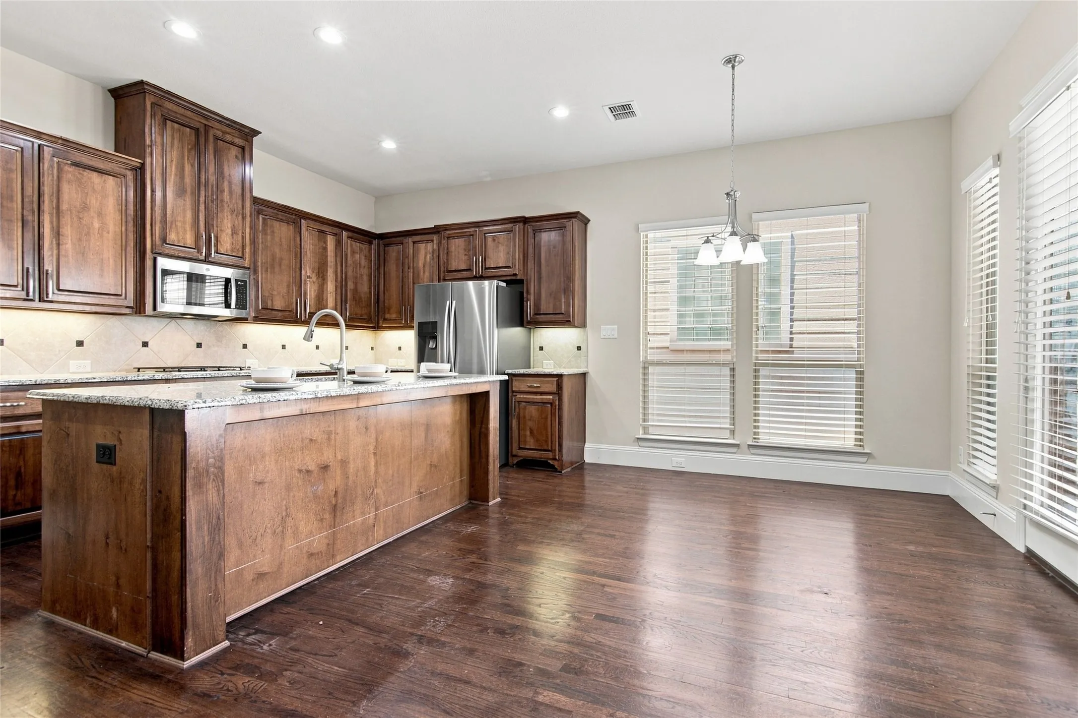 Kitchen featuring wood-finish cabinetry, a large center island with a light-toned countertop, stainless steel appliances, a tiled backsplash, and dark wood-finish flooring