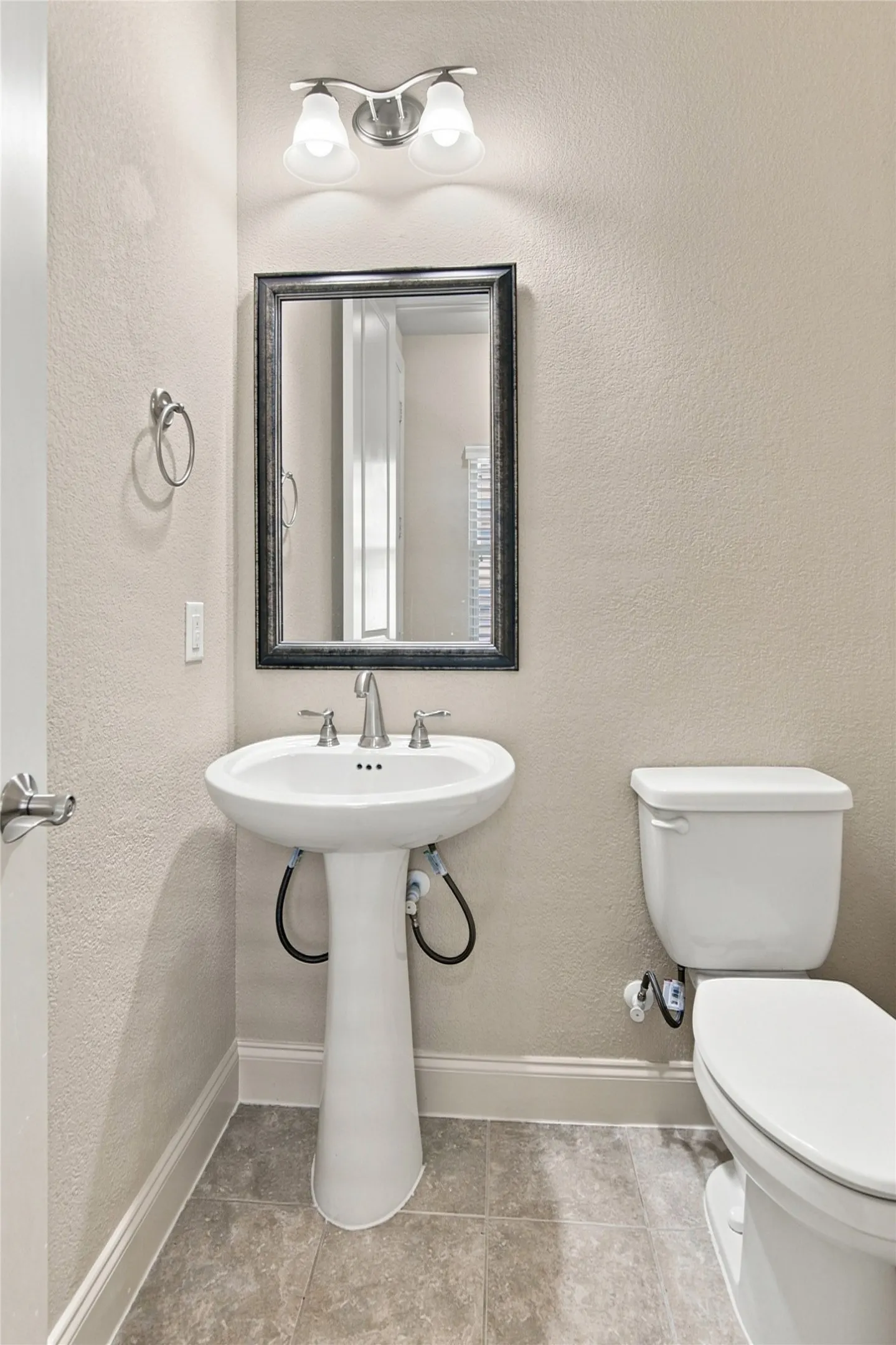 Elegant half bath featuring a classic pedestal sink with chrome widespread faucet, framed mirror with brushed nickel vanity lighting, porcelain toilet, and neutral tile flooring