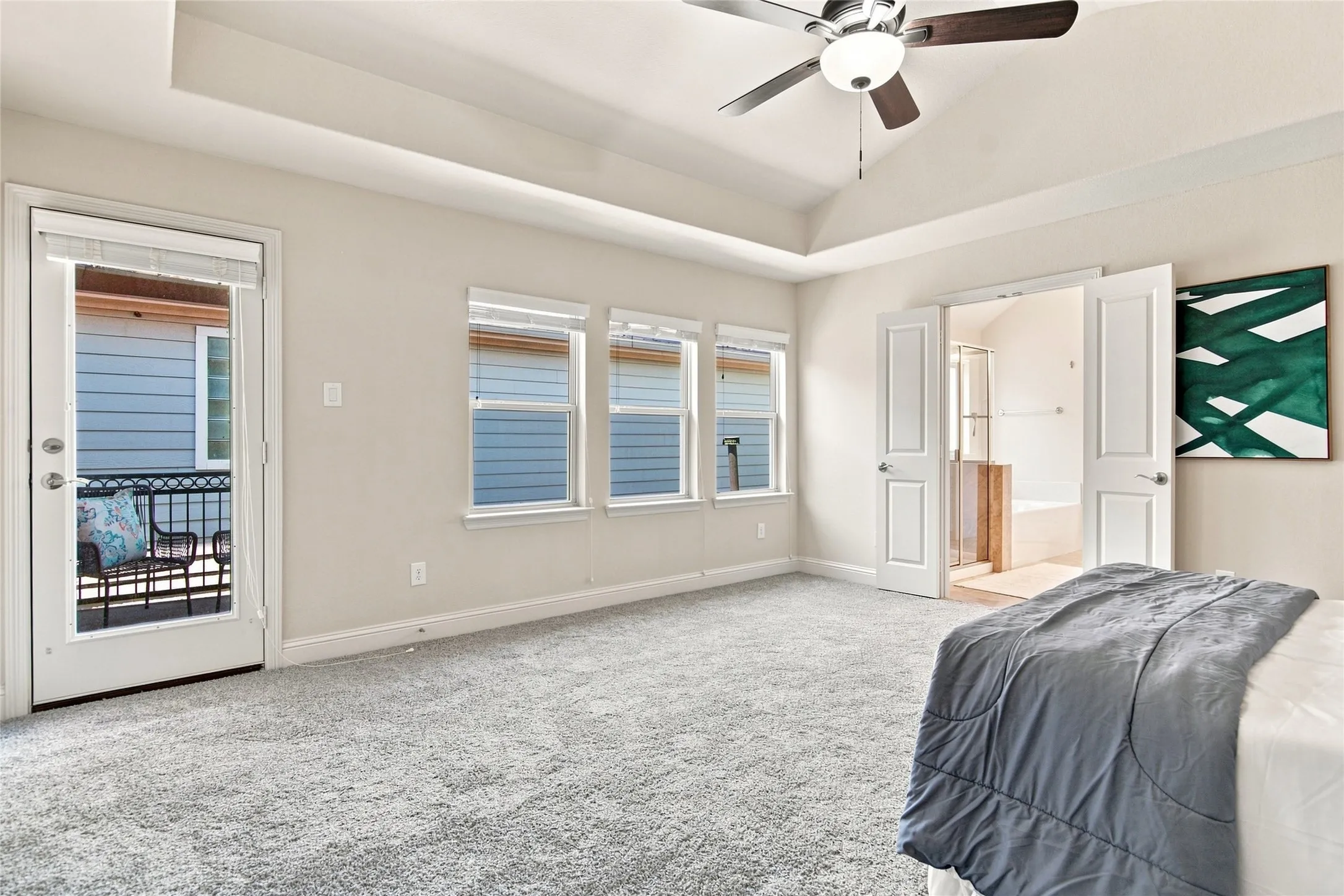 Tray ceiling with a ceiling fan, light gray carpeting, glass-paneled door opening to a balcony with a wrought iron railing, two windows with blinds, and double doors opening to a bathroom with a soaking tub