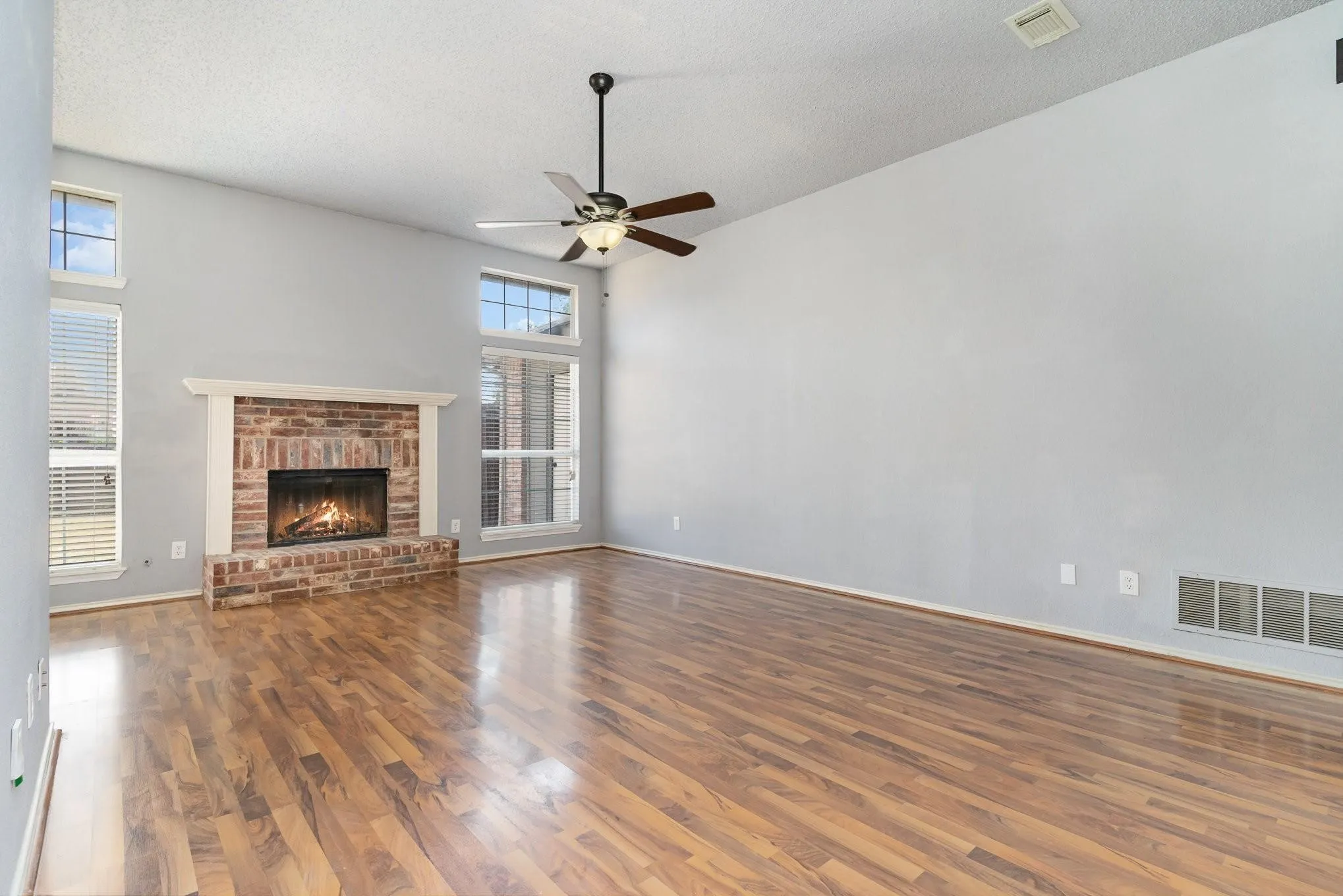Vaulted ceiling with a ceiling fan, brick-surround fireplace with hearth and mantel, wood-finish flooring, and multiple windows