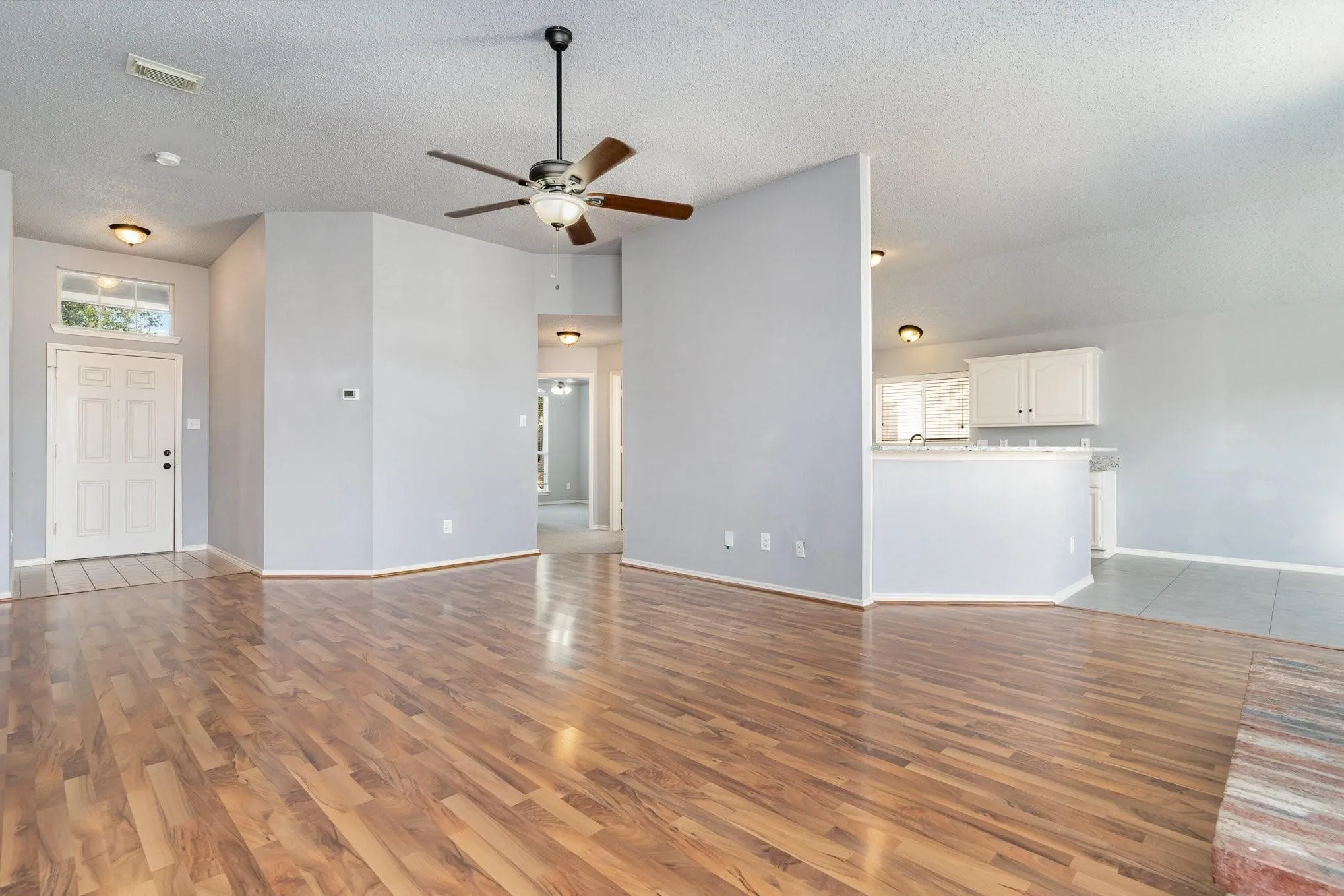Wood-finish flooring throughout, ceiling fan with integrated lighting, white paneled entry door, kitchen pass-through with white cabinetry, and recessed lighting