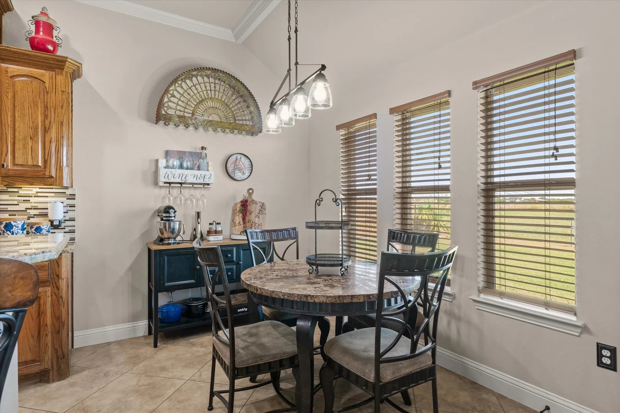 Vaulted ceiling with crown molding, natural wood-finish cabinetry, tile flooring, textured backsplash, and multiple windows with horizontal blinds