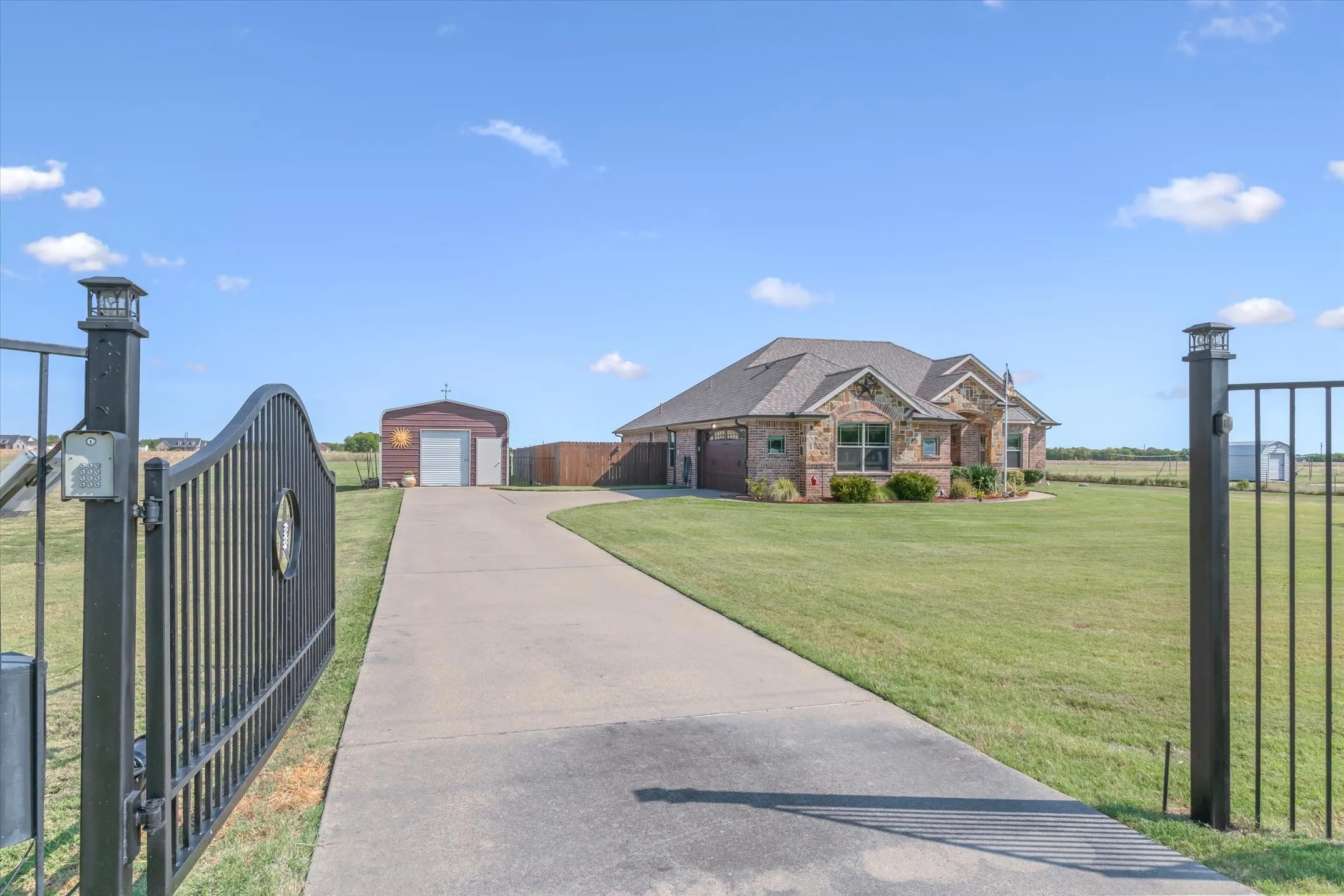 Gated property entrance with a paved driveway, expansive lawn, and a stone-accented exterior