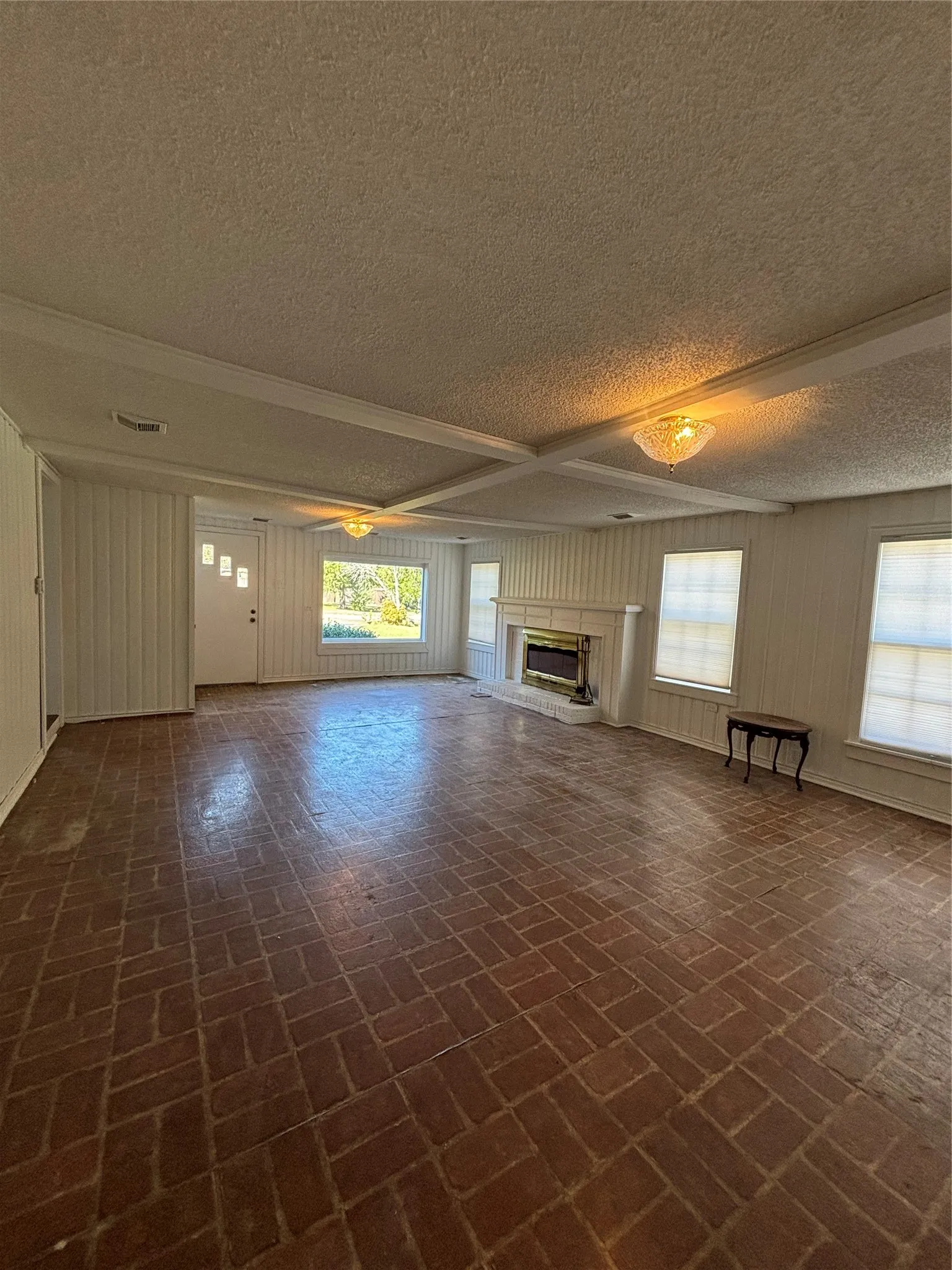 Unfurnished living room with a textured ceiling, a glass covered fireplace, beamed ceiling, and brick patterned flooring