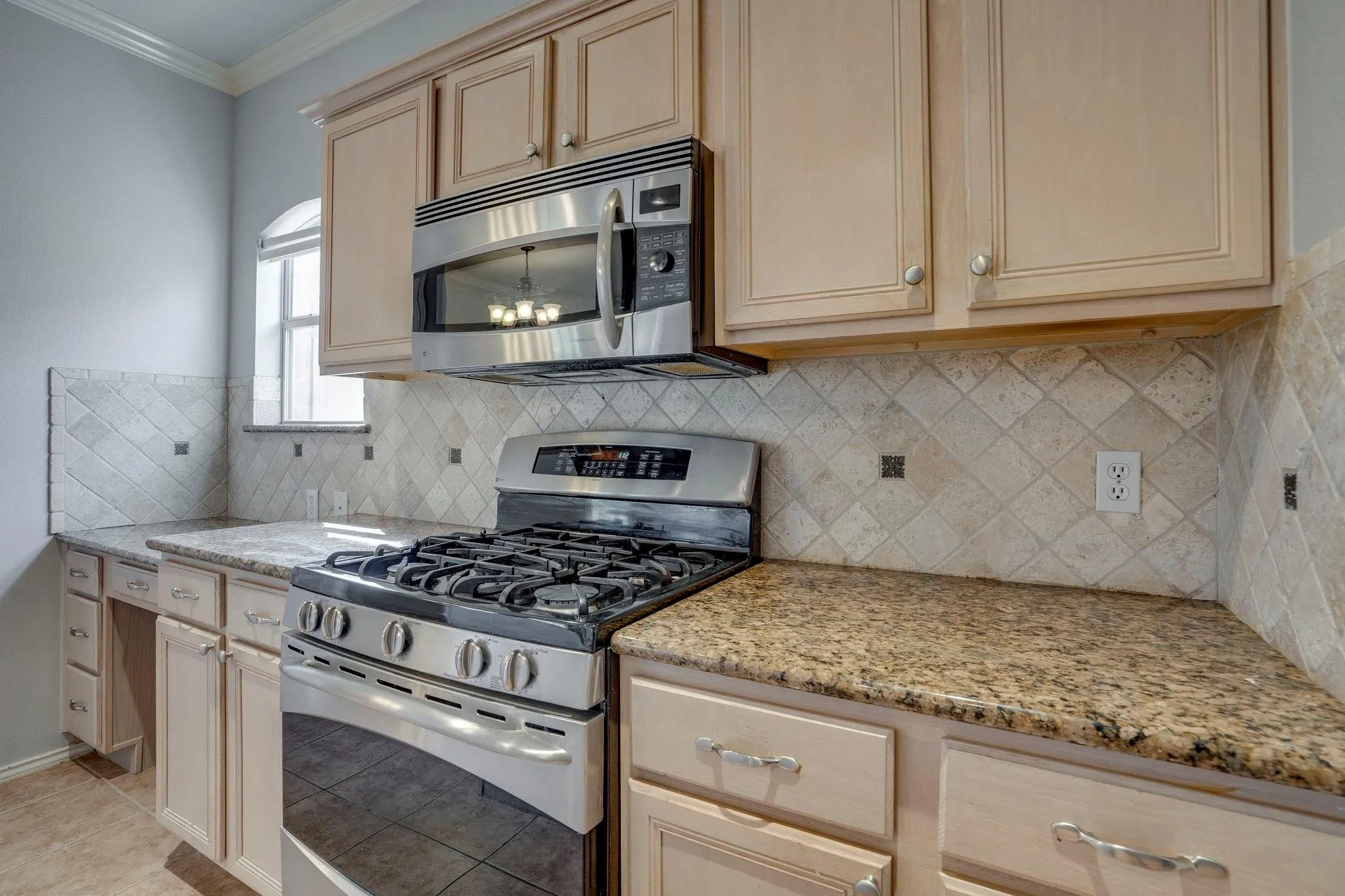 Kitchen featuring light-toned cabinetry, a built-in stainless steel microwave, a gas range, granite-style countertops, and a patterned tile backsplash
