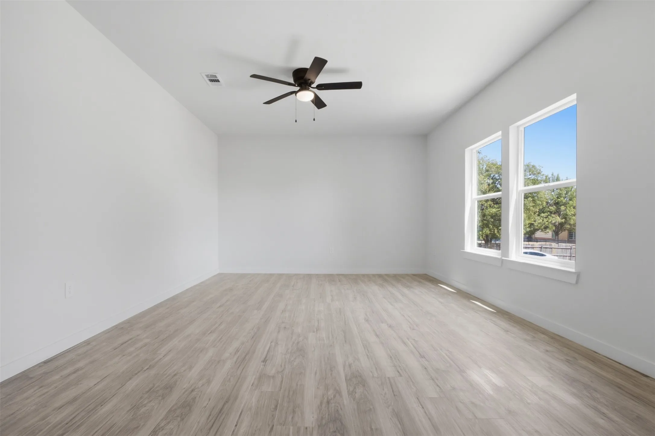 Light wood-finish flooring, two large windows, and a ceiling fan with integrated lighting