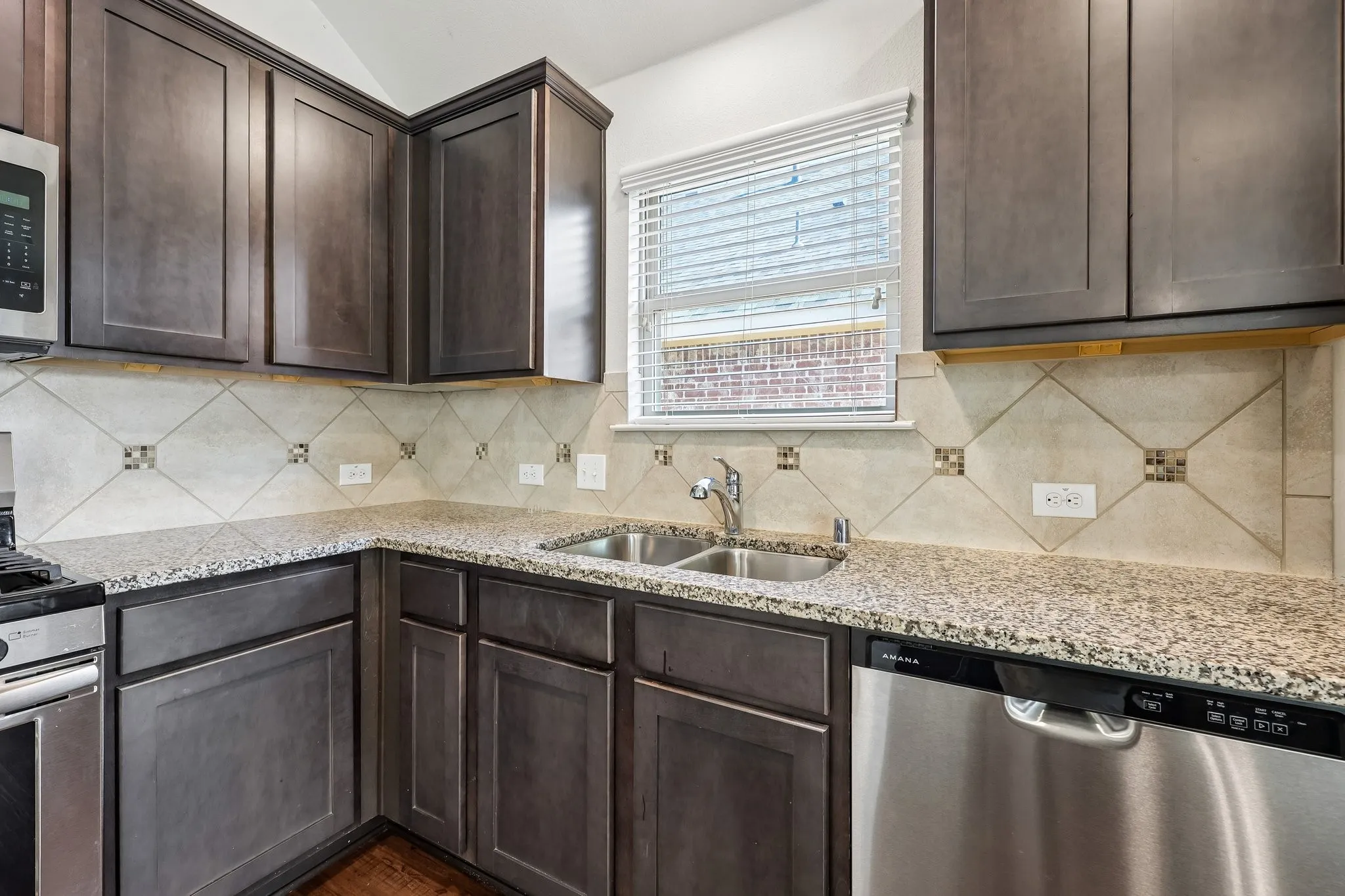 Kitchen featuring dark wood-finish cabinetry, speckled countertops, tile backsplash with decorative accents, stainless steel double basin sink, and stainless steel dishwasher