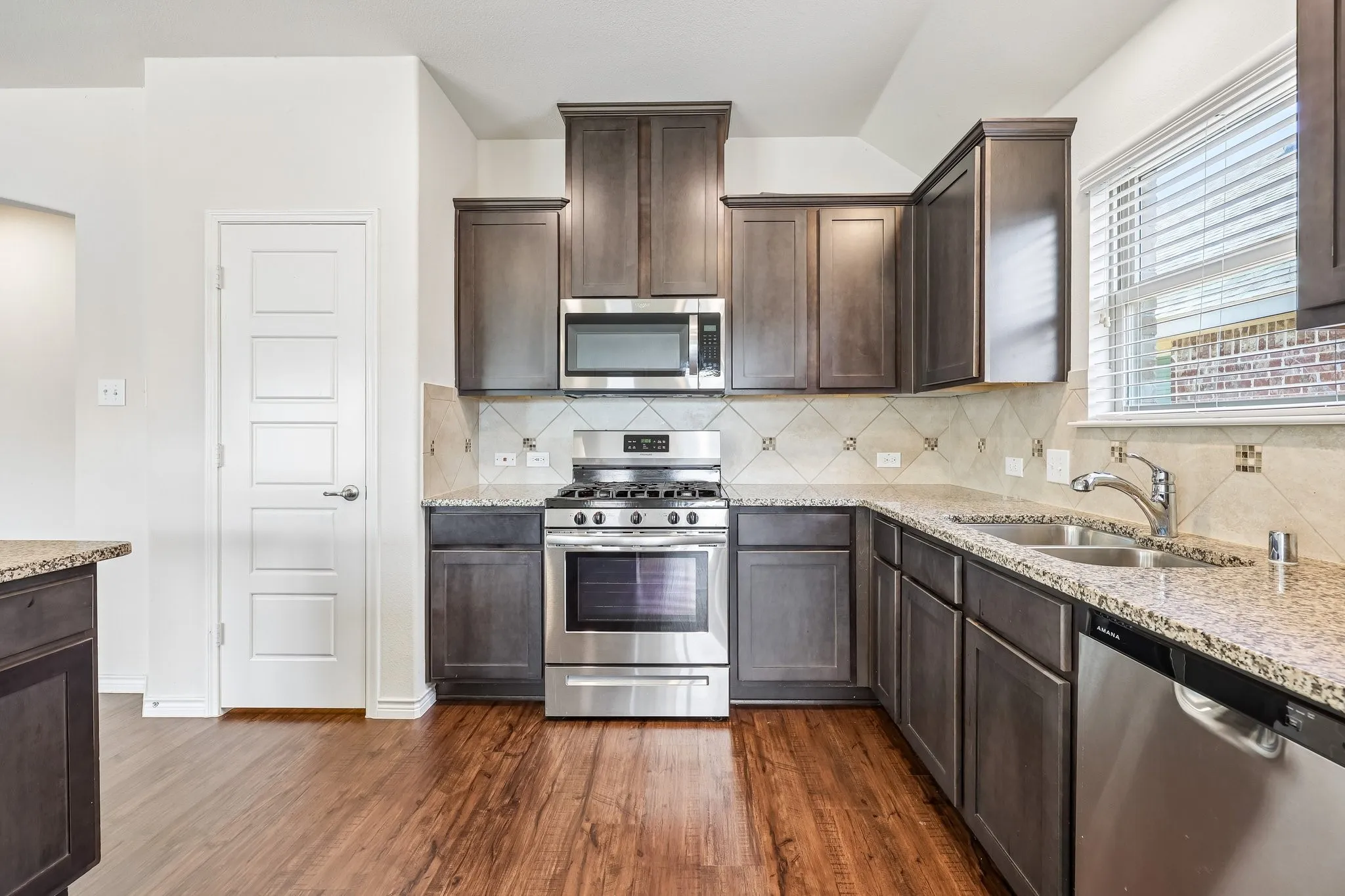 Wood-finish flooring with dark wood cabinetry, stainless steel appliances, speckled countertops, and a tiled backsplash