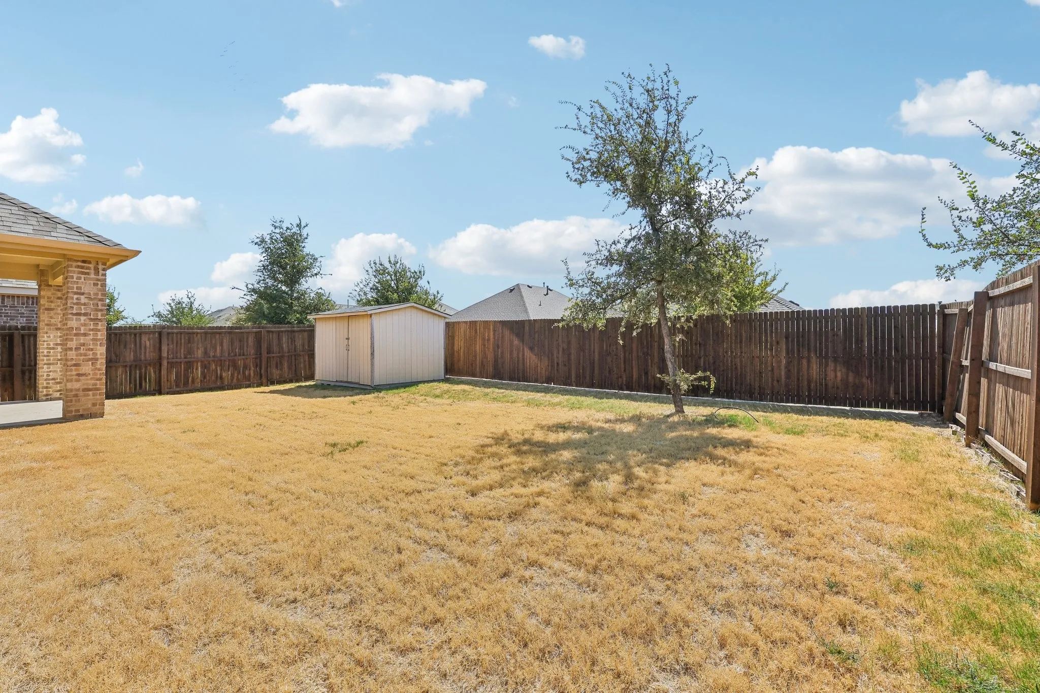 Expansive backyard featuring a privacy fence, covered patio with brick columns, a mature tree, and a storage shed