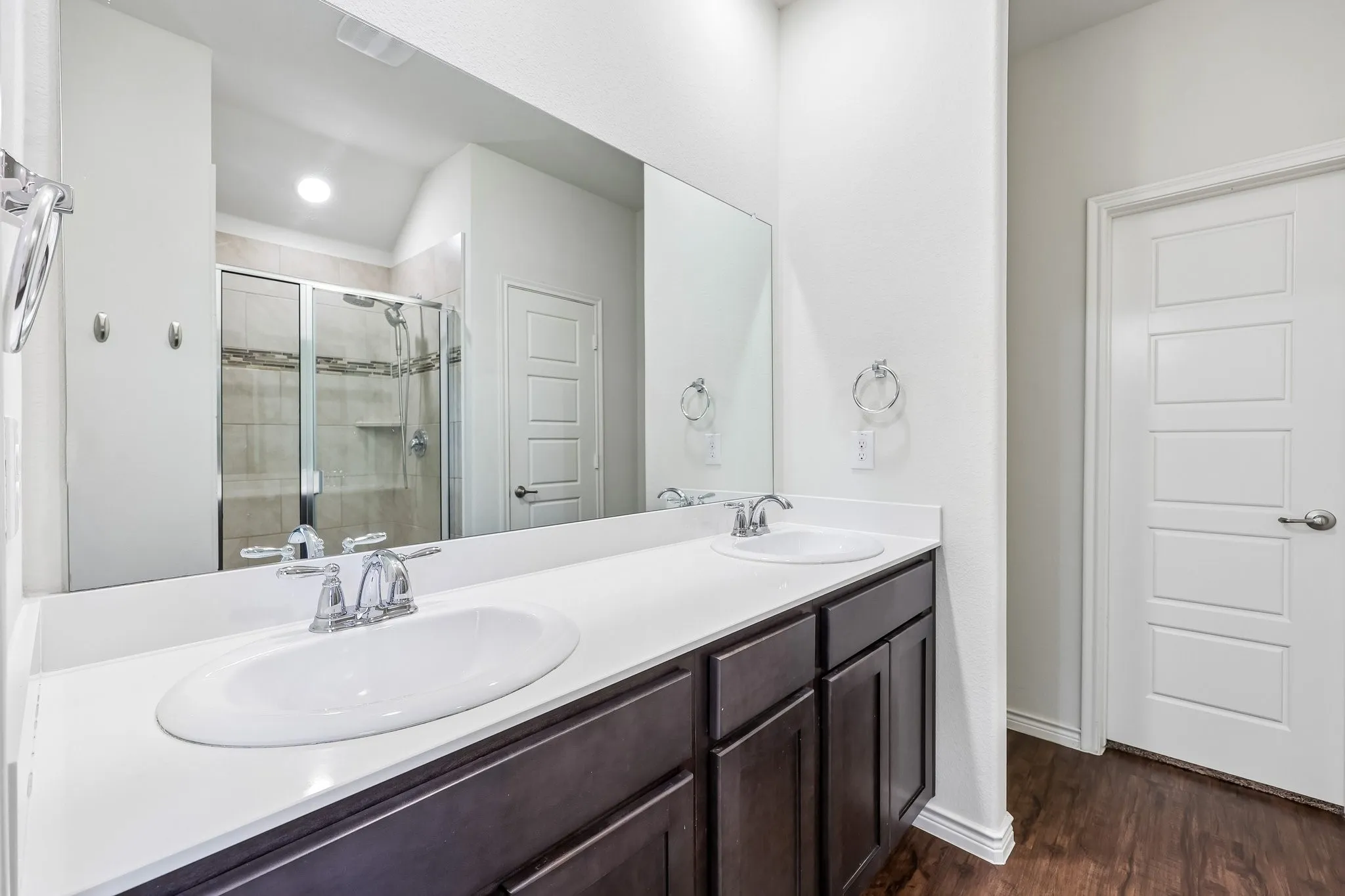 Dual undermount sinks with chrome fixtures, a spacious countertop, dark wood-finish cabinetry, a tiled walk-in shower with glass enclosure, and wood-finish flooring