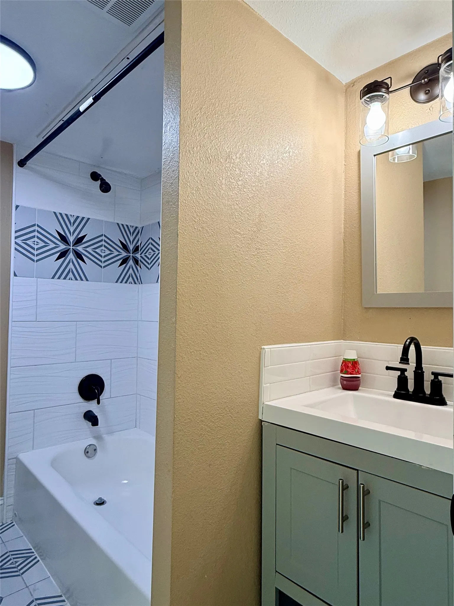 White bathtub with matte black fixtures, subway tile surround with decorative accent, and patterned floor tiles