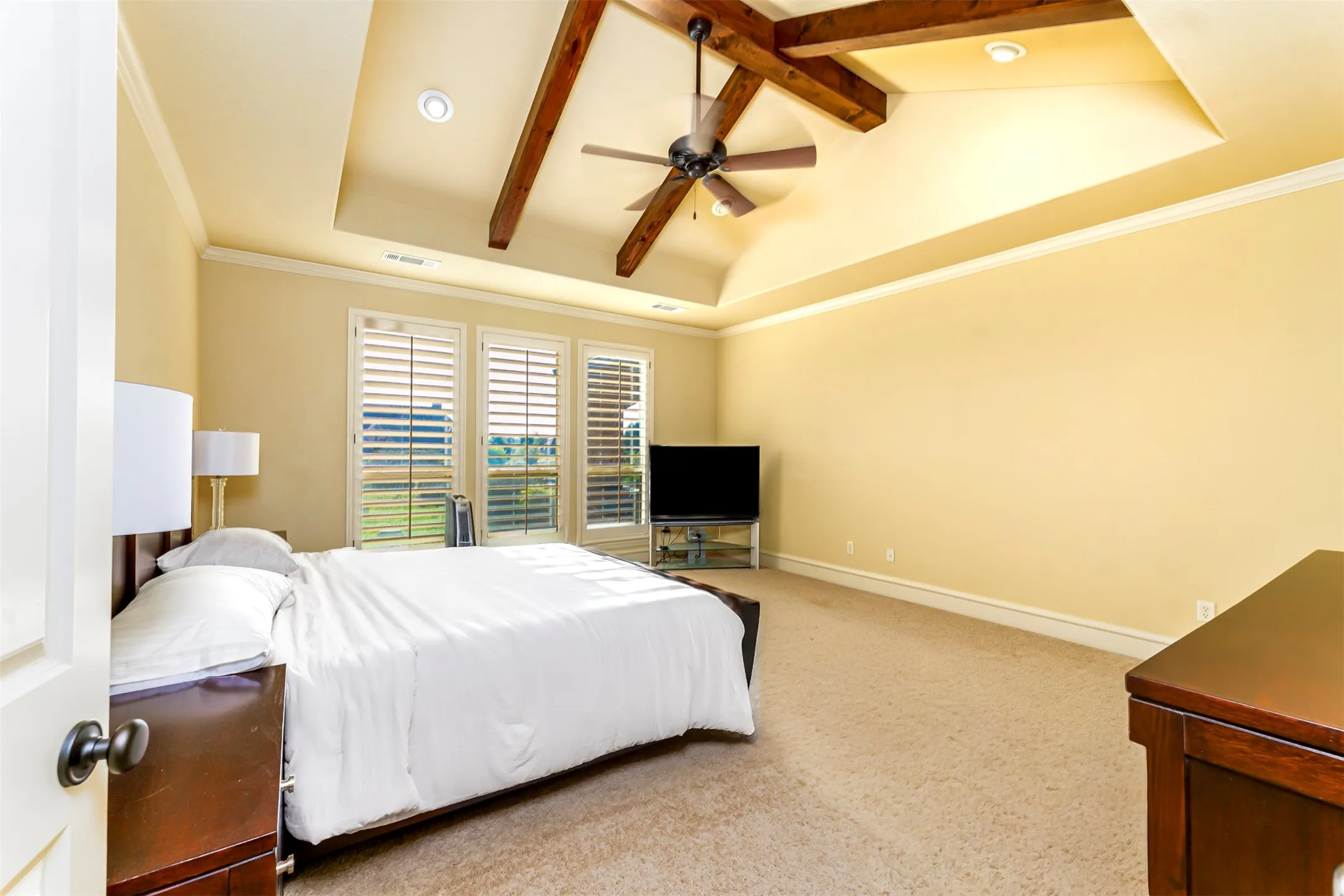 Tray ceiling with exposed wood beams, ceiling fan, recessed lighting, carpet flooring, and plantation shutters