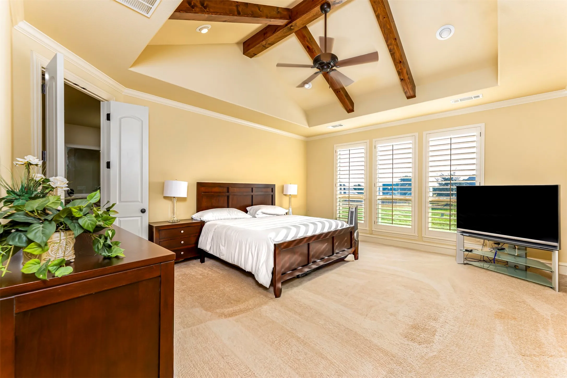 Tray ceiling with exposed wood beams, ceiling fan, recessed lighting, and three windows with plantation shutters