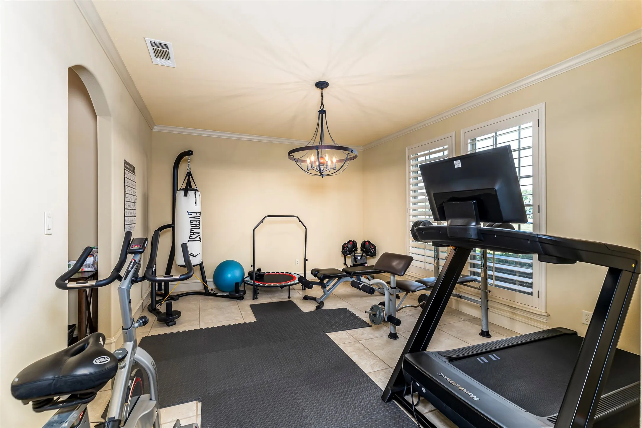 Spacious room with tile flooring, dual windows featuring plantation shutters, crown molding, an arched entryway, and a decorative chandelier