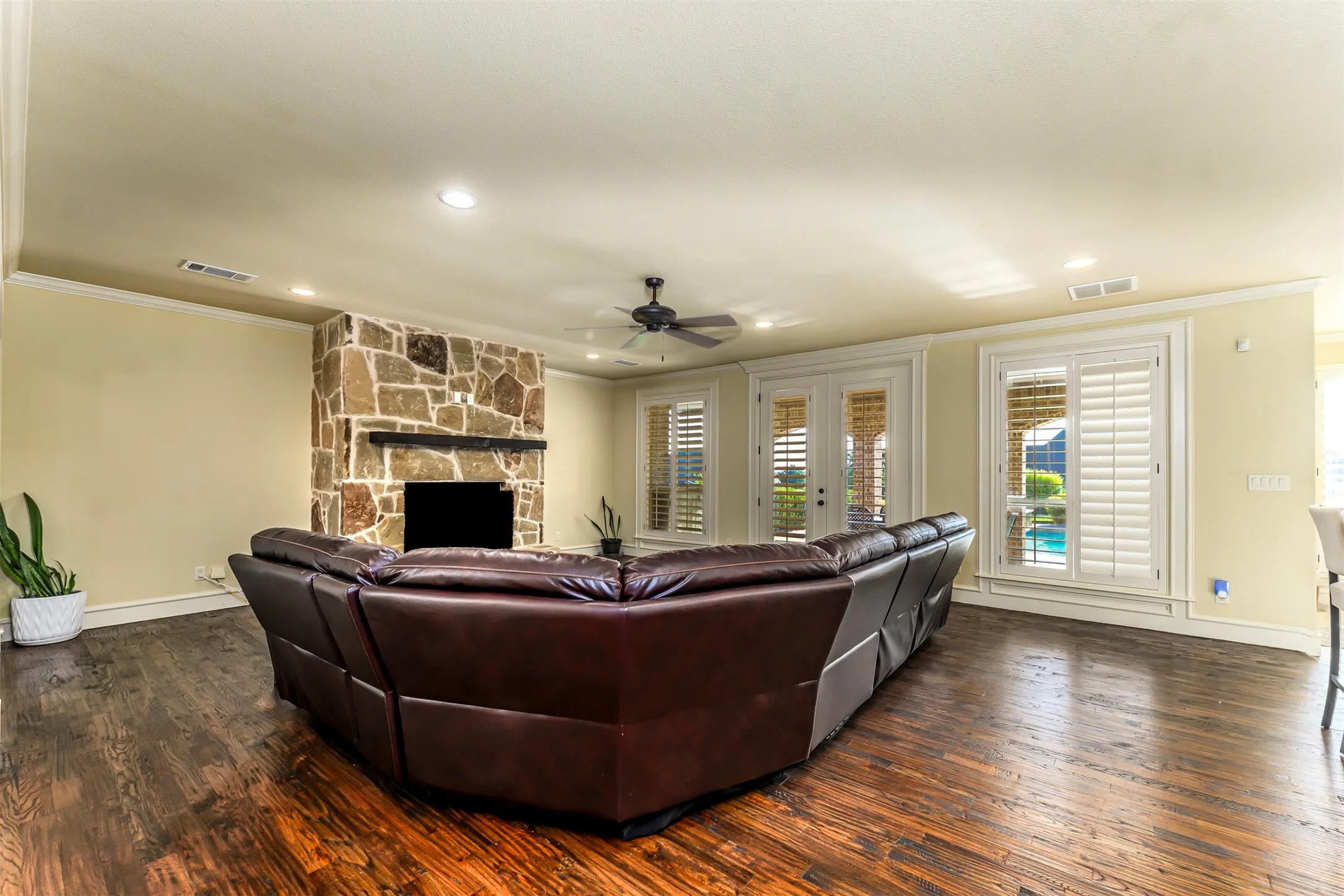 Stone-clad fireplace with mantel, dark wood-finish flooring, recessed lighting, ceiling fan, and French doors with plantation shutters