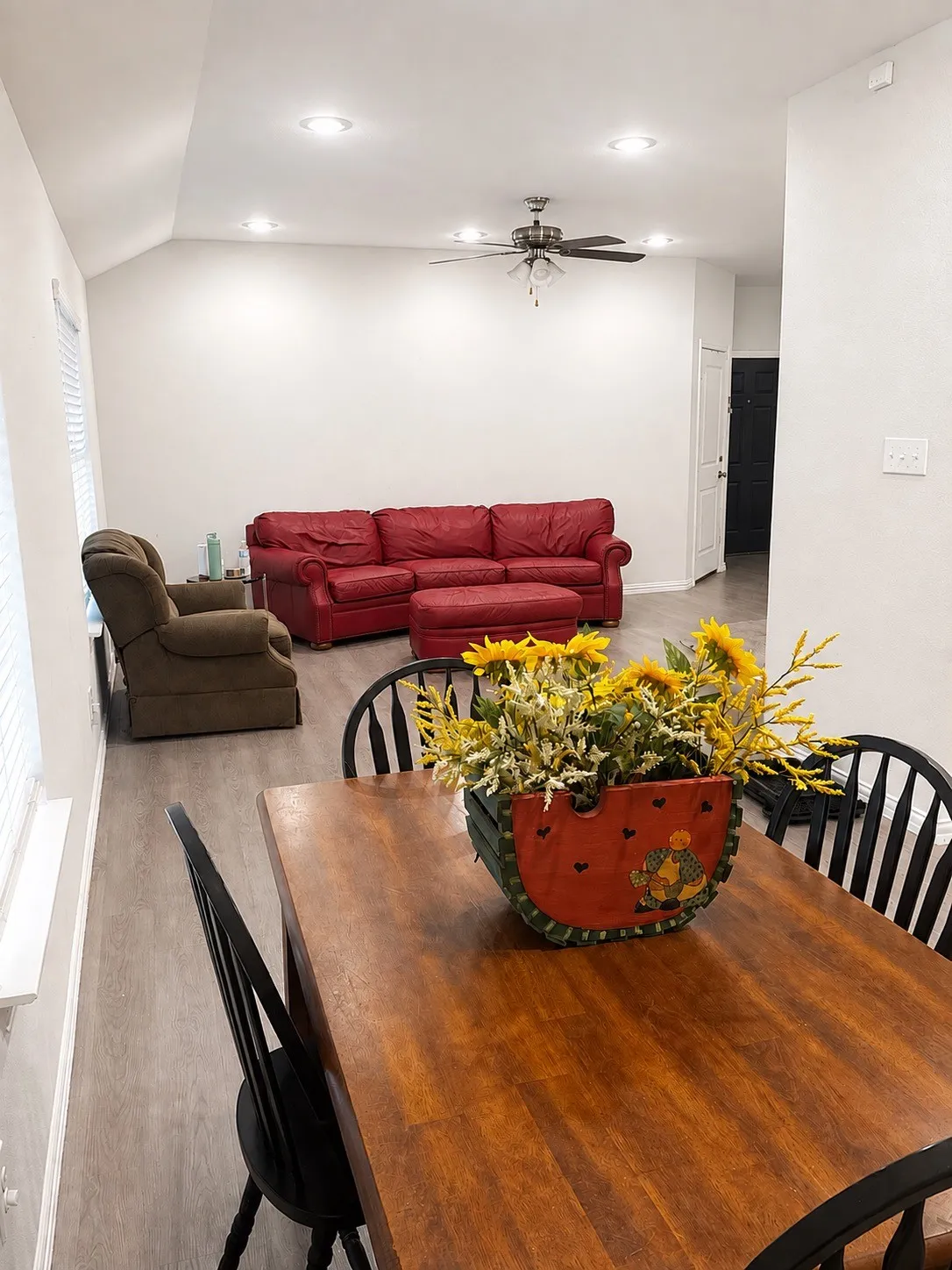 Open-concept living area featuring wood-finish dining table with seating for four, recessed ceiling lighting, ceiling fan with integrated light fixture, and direct sightlines to the adjoining living room with red upholstered sofa and brown upholstered armchair