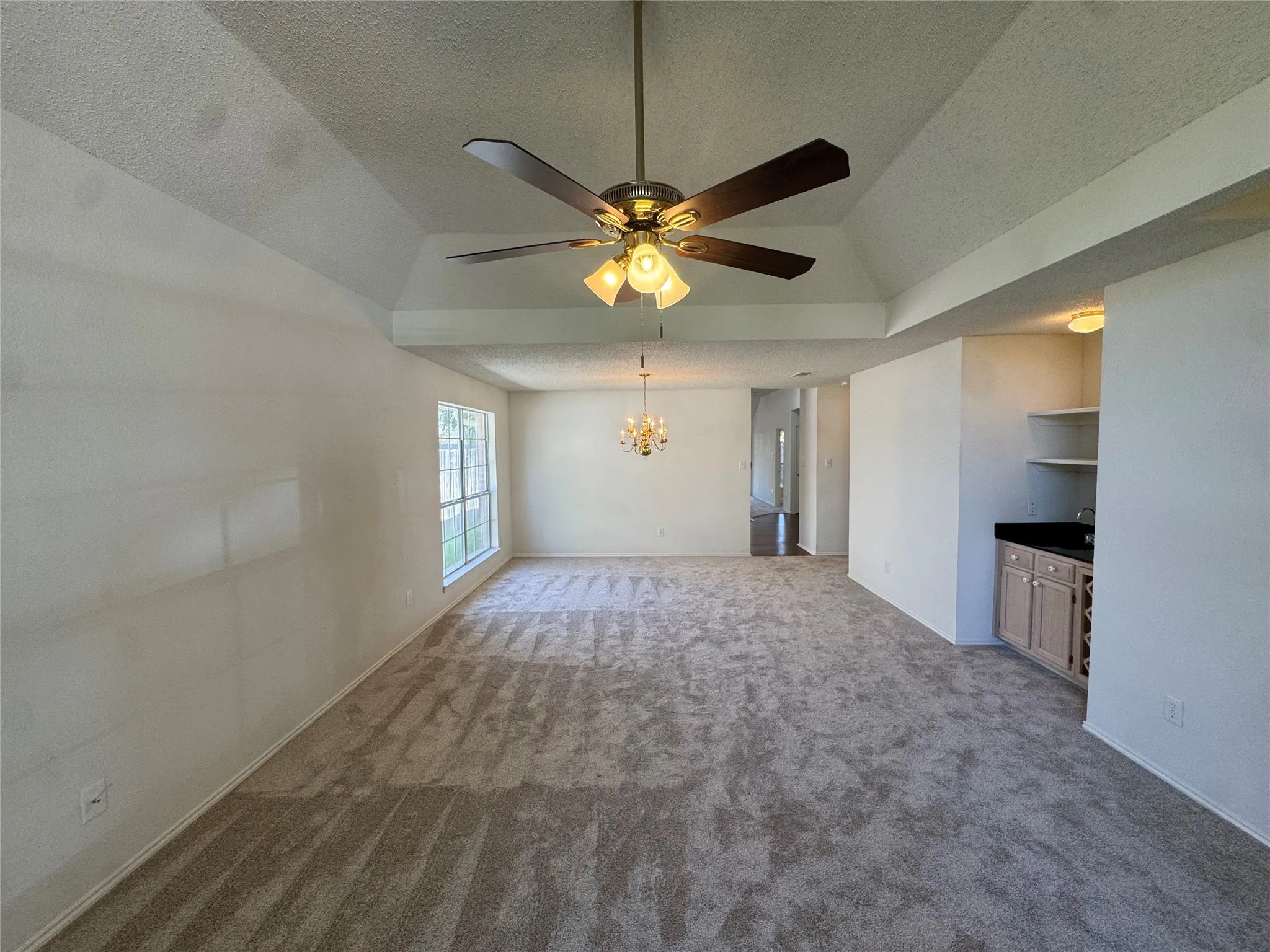 Spacious room featuring a tray ceiling, ceiling fan with integrated lighting, carpeted flooring, and a built-in wet bar with cabinetry, shelving, and a dark countertop