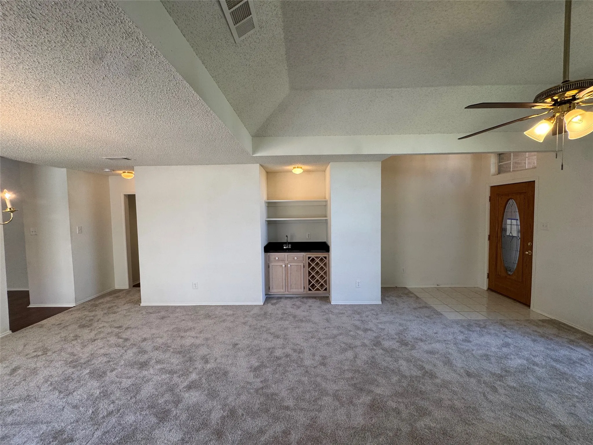 Expansive carpeted living area with a vaulted ceiling, built-in wet bar with shelving, wood-finish front door with an oval glass insert, and a ceiling fan with integrated lighting