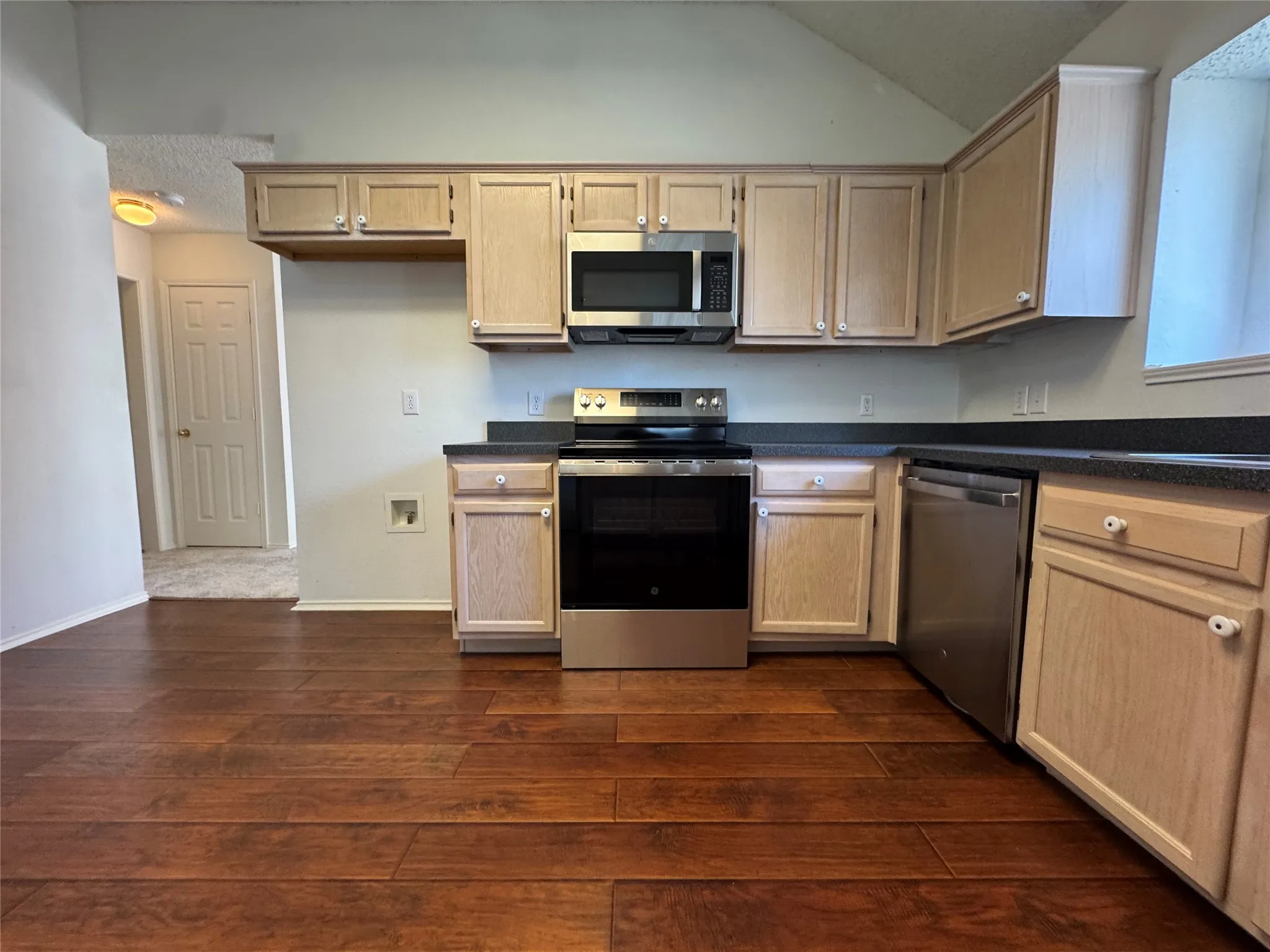 Kitchen featuring wood-finish flooring, stainless steel appliances, light wood cabinetry, and dark countertops