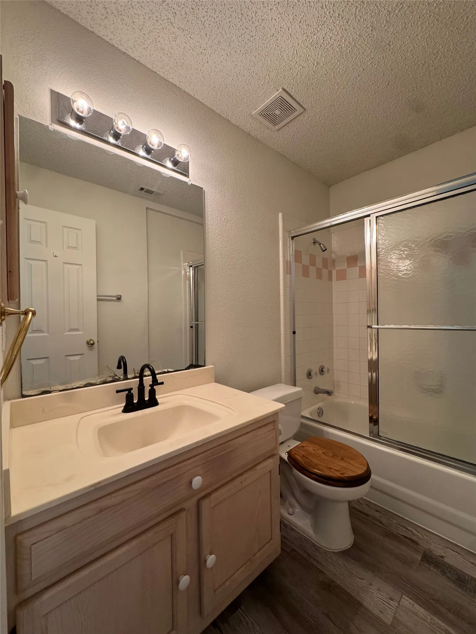 Wood-finish flooring, light-toned vanity with integrated sink, chrome vanity lighting, and a bathtub with sliding shower doors