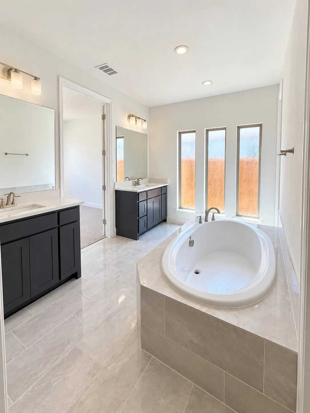 Freestanding soaking tub with tiled surround, dual vanity sinks with dark cabinetry, large mirrors, and recessed lighting