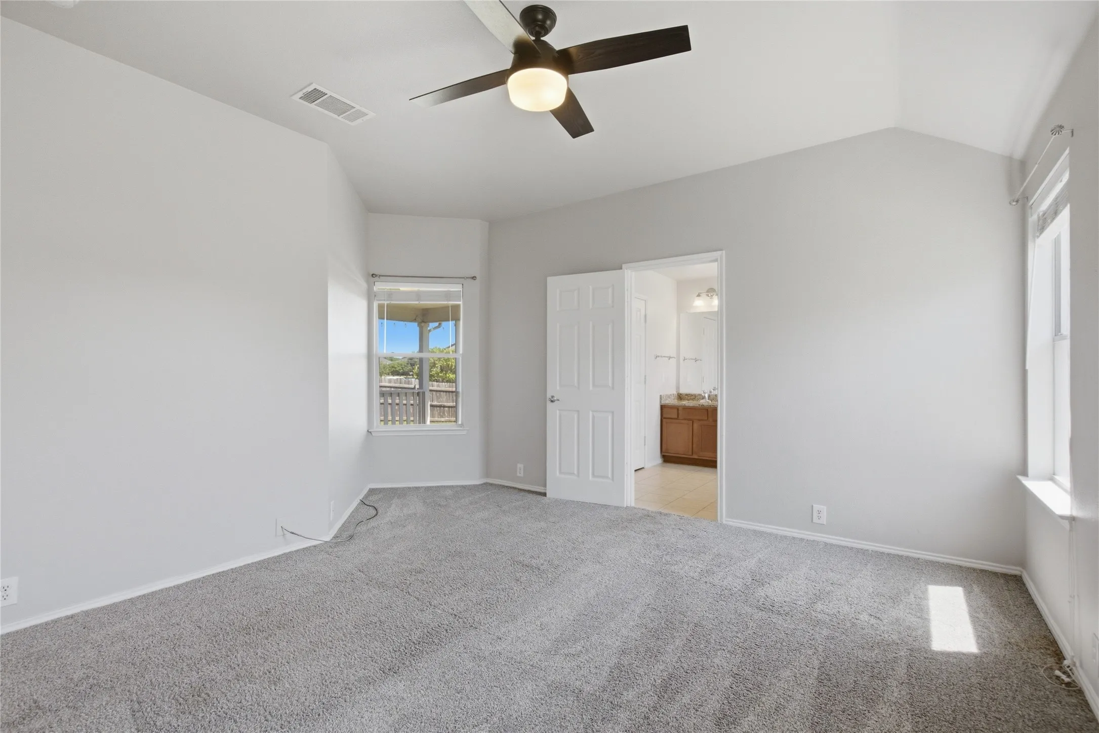 Vaulted ceiling with an attached ceiling fan, two windows providing natural light, and plush gray carpeting