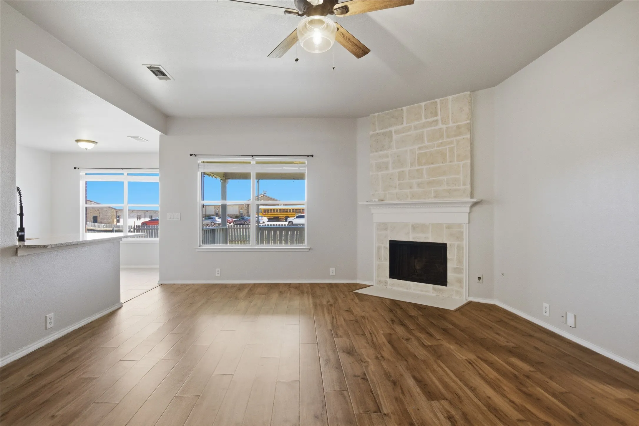 Wood-finish flooring, stone-clad fireplace, ceiling fan with light fixture, large windows, and a breakfast bar with a sink