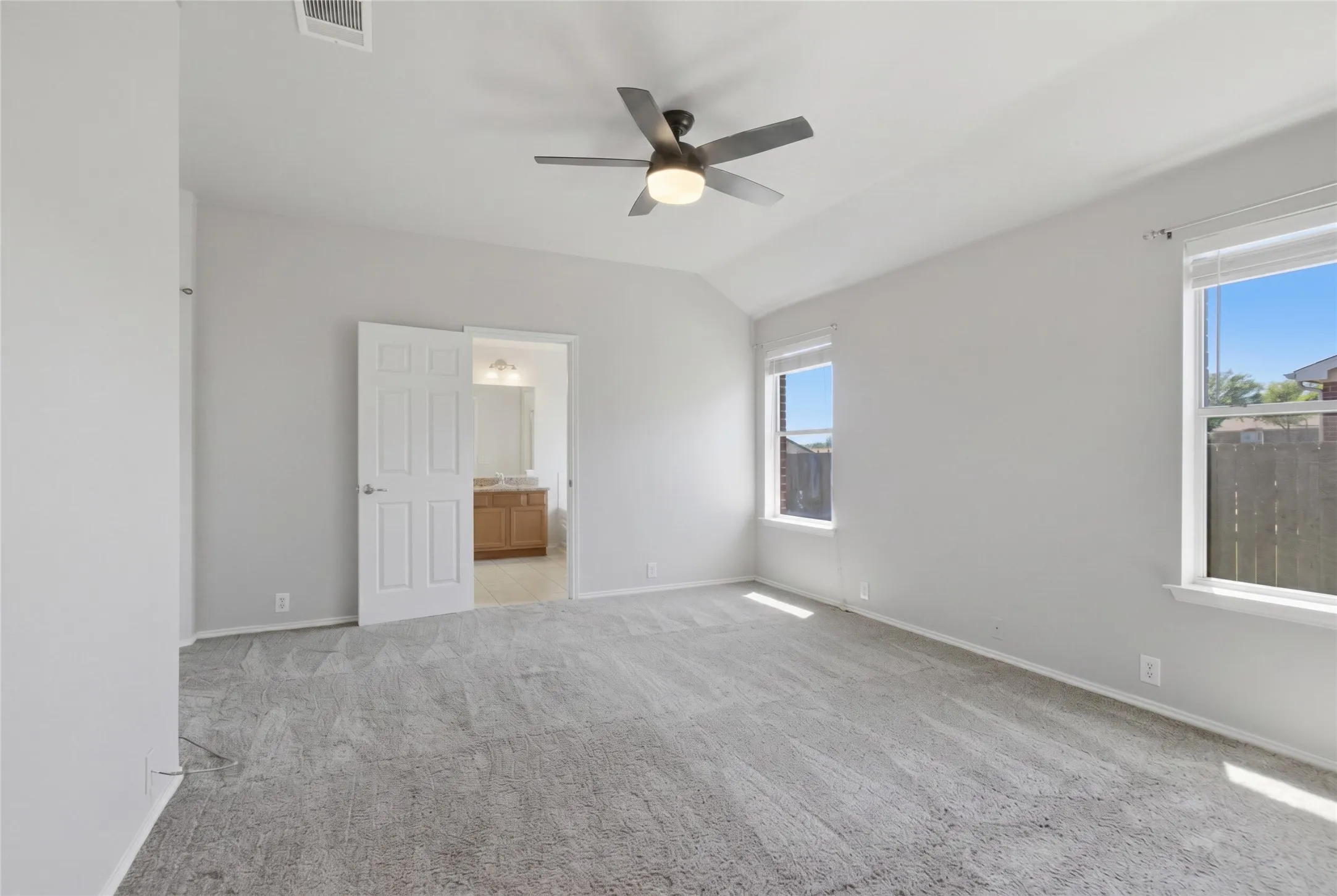 Vaulted ceiling, two windows, ceiling fan with integrated lighting, gray carpet flooring, and a white paneled door