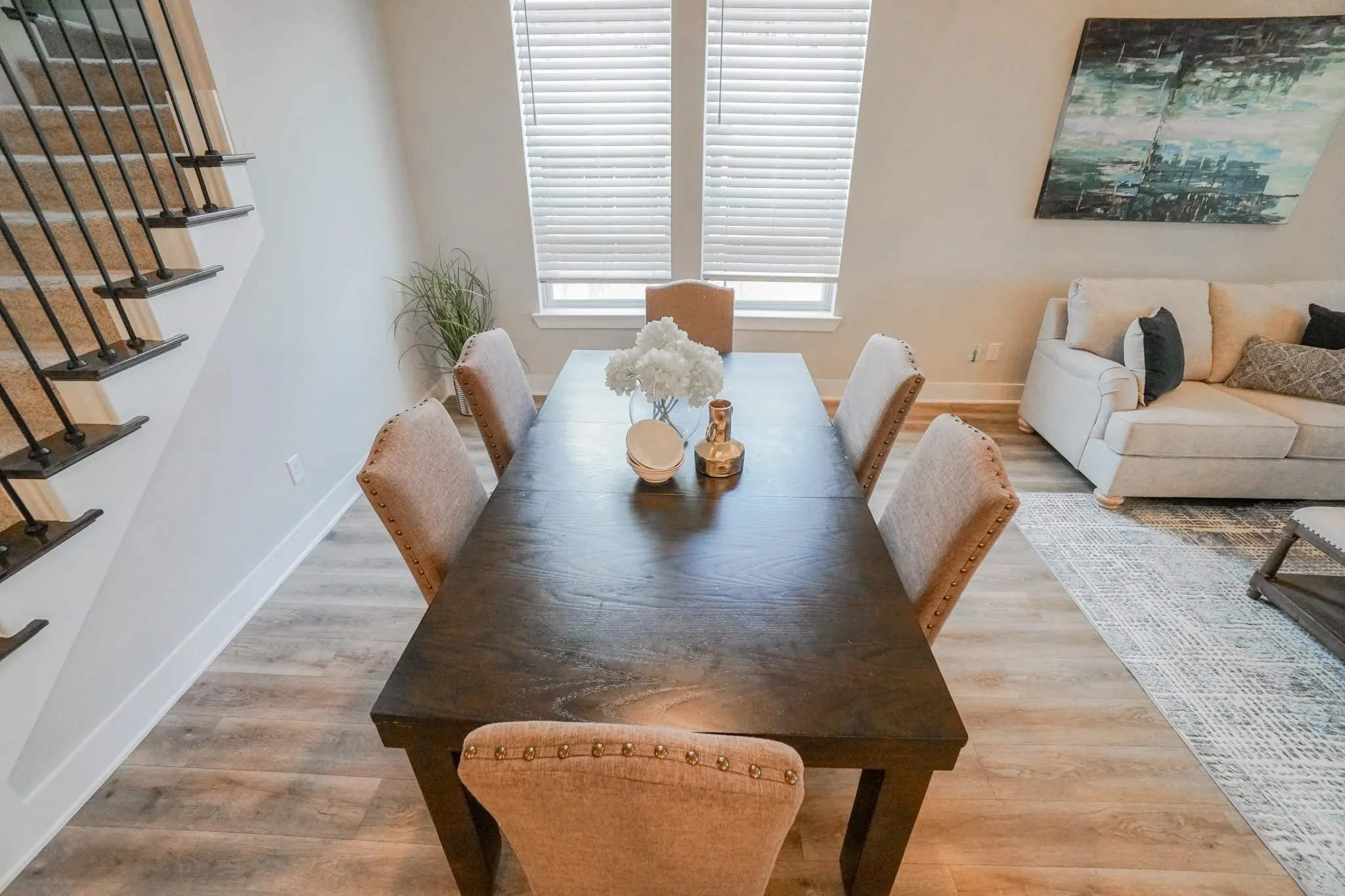 Wood-finish flooring throughout, bright window with blinds, and a staircase featuring wrought iron balusters