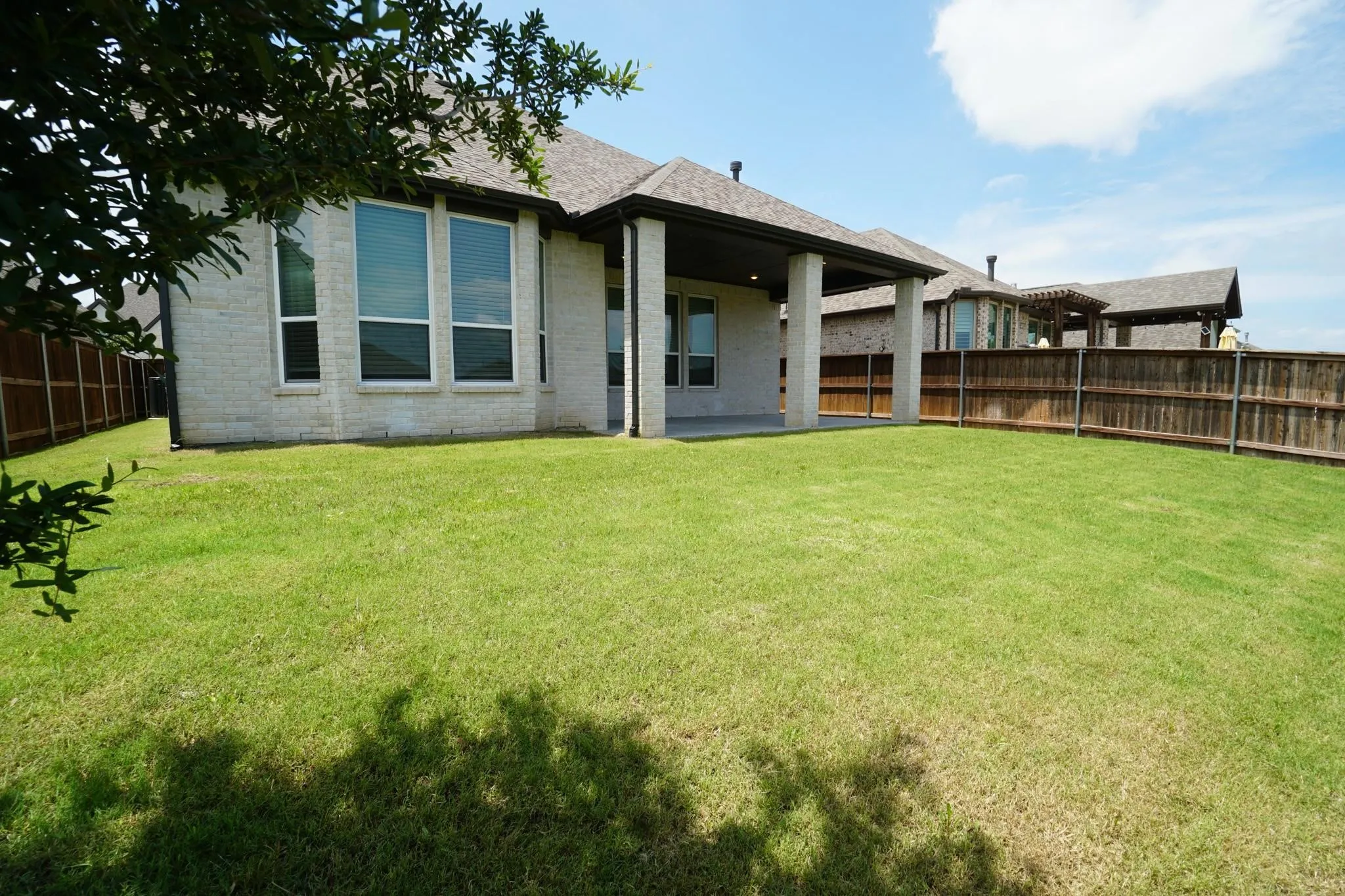Expansive grassy yard featuring a covered patio, light brick exterior, dark wood fencing, and multiple windows