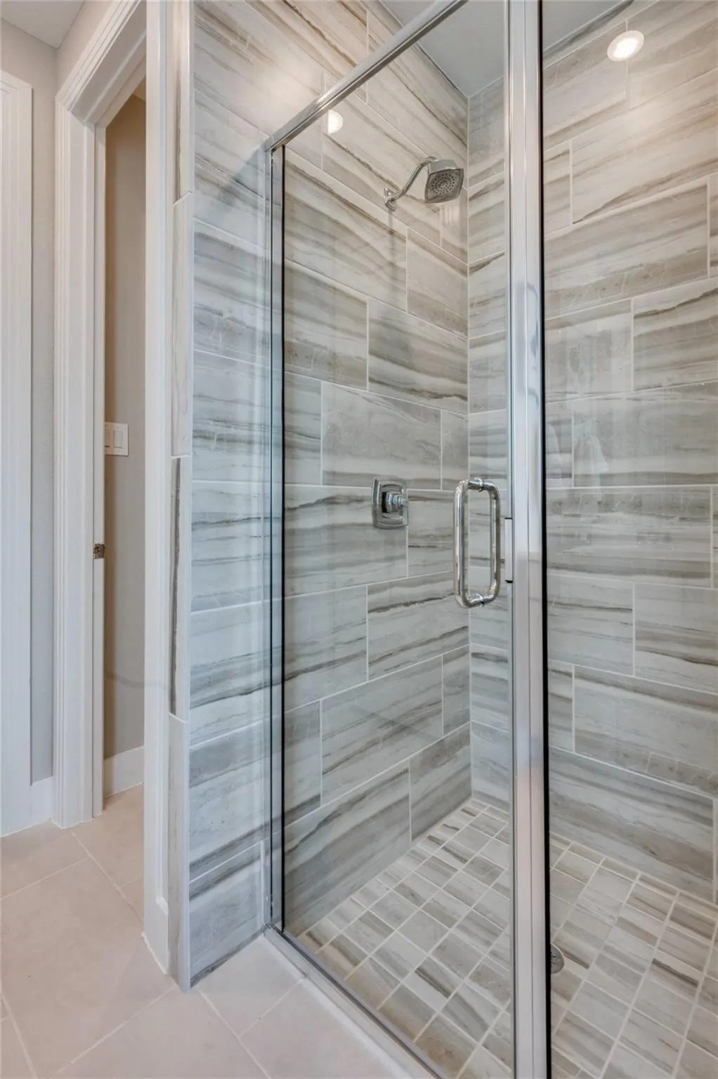 Glass-enclosed shower with a chrome shower head and handle, featuring large format wall tiling and a tiled shower pan