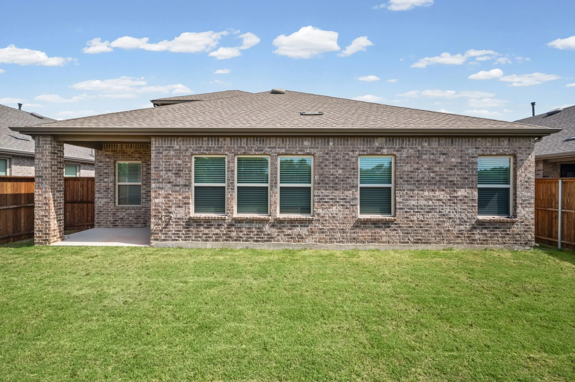Brick exterior with a covered patio, shingle roof, and a grassy lawn