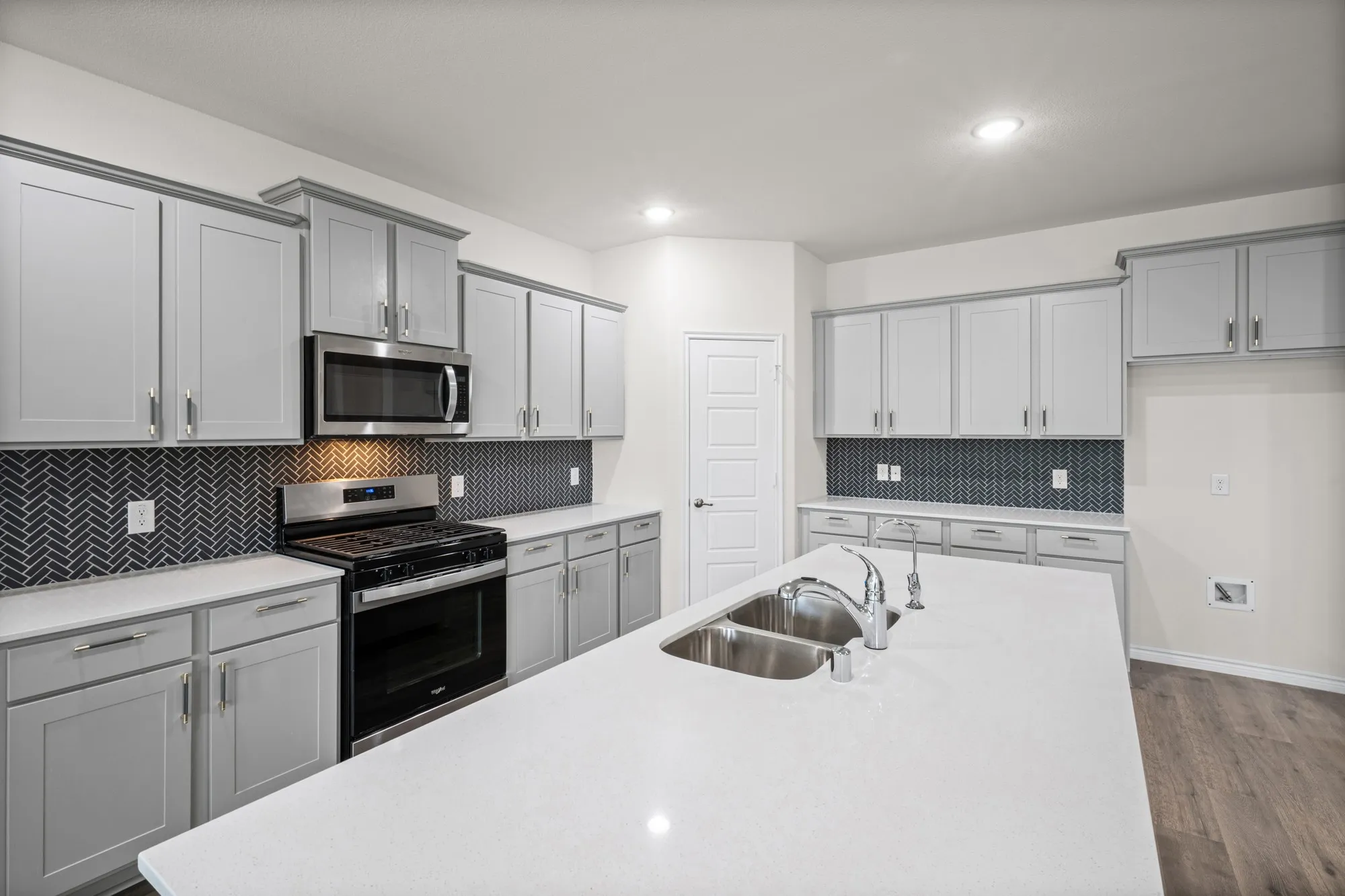 Kitchen featuring an expansive island with a double basin sink and chrome fixtures, light gray cabinetry with crown molding, stainless steel range and microwave, and a dark herringbone tile backsplash