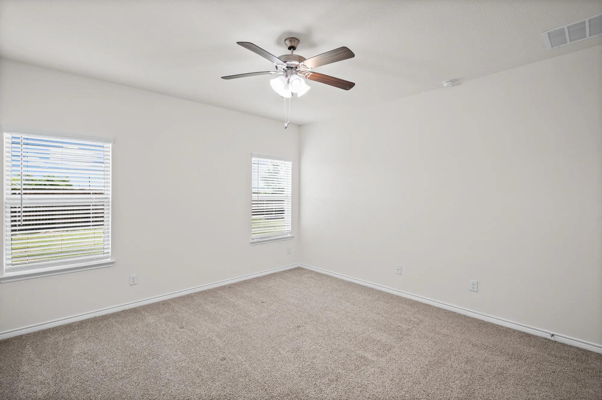 Neutral wall paint, carpet flooring, two windows with blinds, and a ceiling fan with light fixture