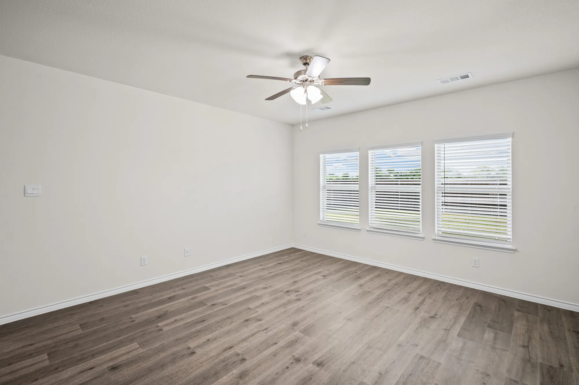 Spacious room featuring wood-finish flooring, three windows with blinds, a ceiling fan with light fixture, and neutral wall tones